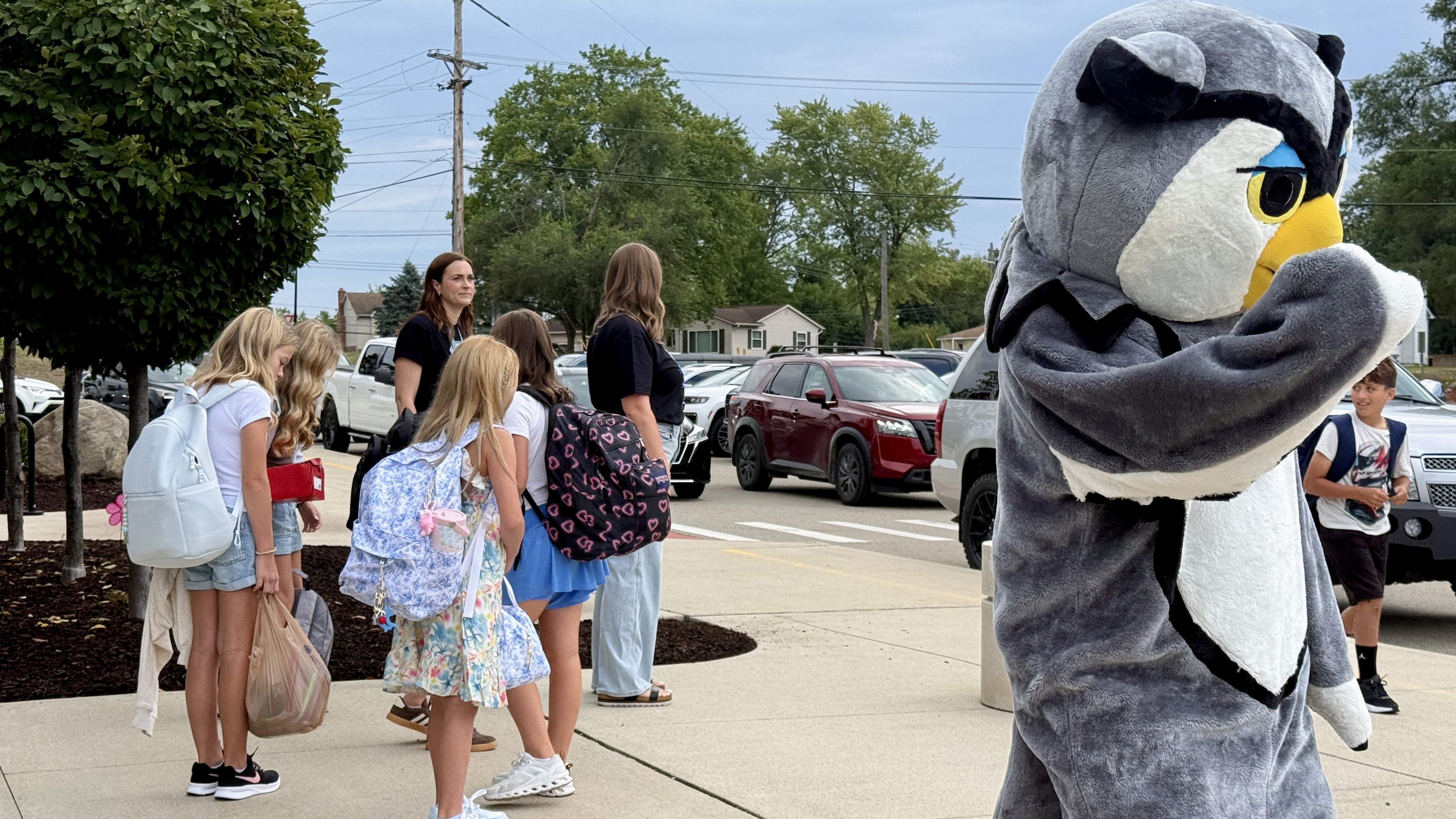 Photo of a person in an Owl costume waving at people with several kids gathered on a sidewalk nearby