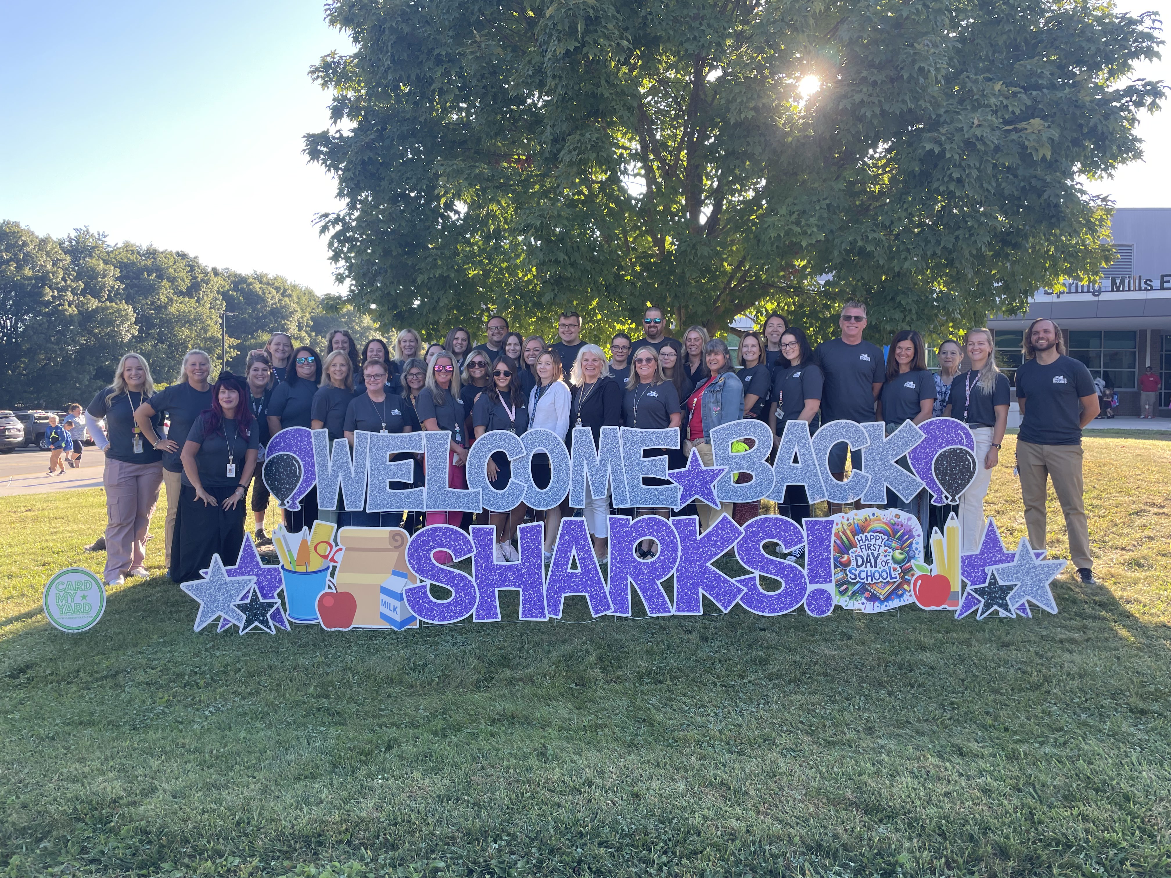 Staff posing in front of a sign in the grass that reads "spring mills sharks"