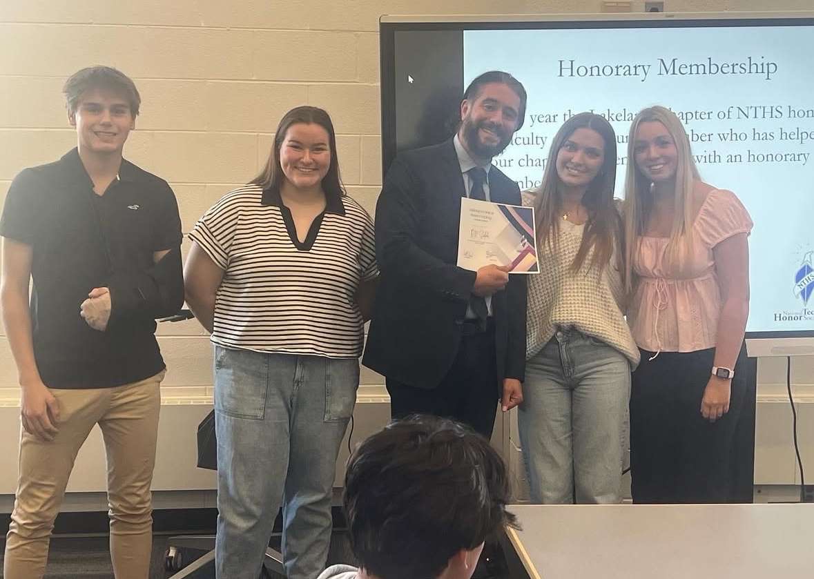 5 people standing in front of a screen giving an award to the one in the middle