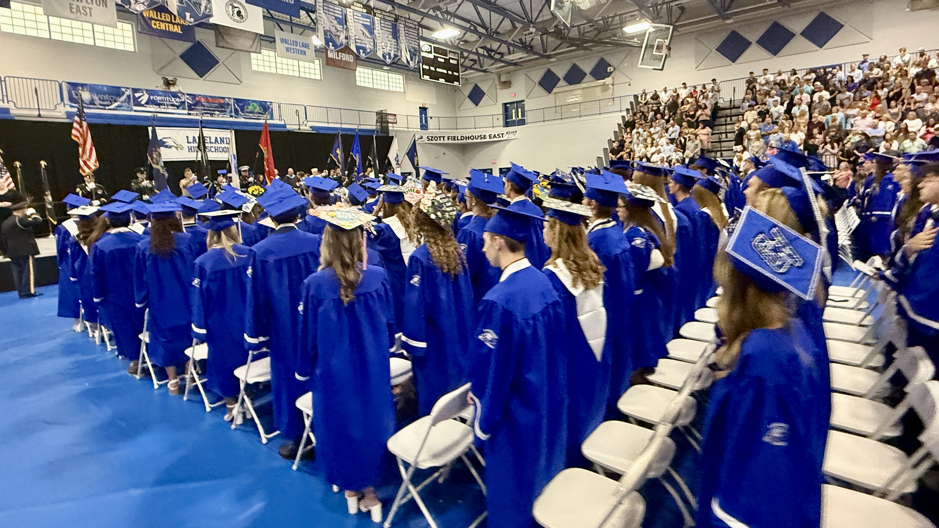 STUDENTS IN BLUE CAPS AND GOWNS AT GRAD CEREMONY IN GYM