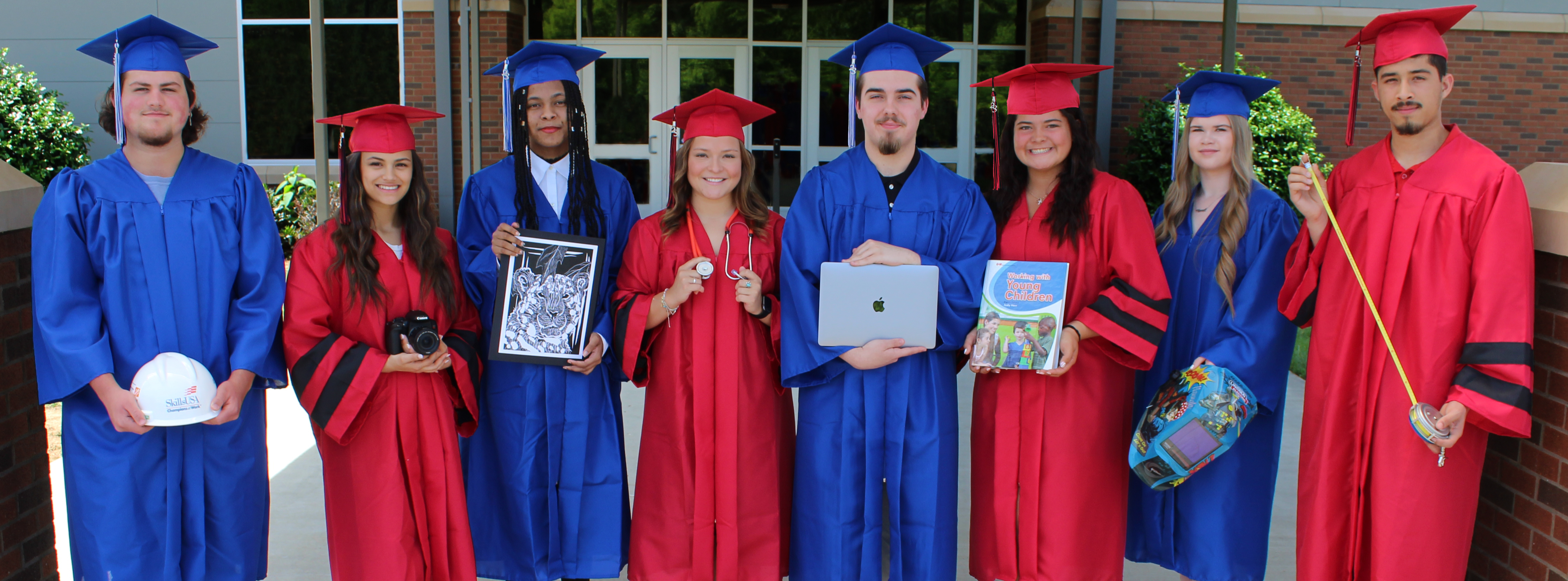 A group of graduating seniors wear their cap and gowns and hold items they will use in their future careers