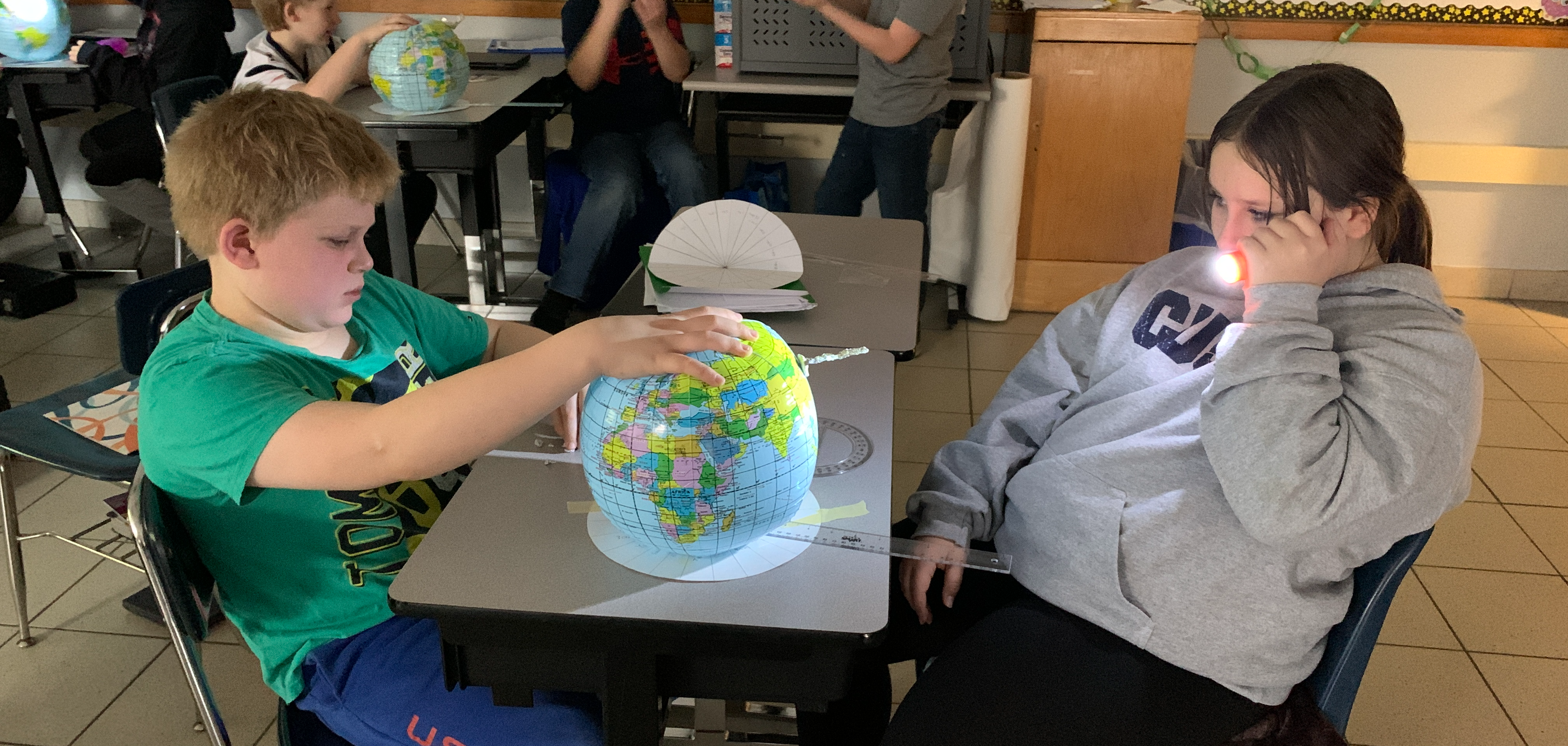 two students participate in a science experiment using a flashlight and a mock model of the earth