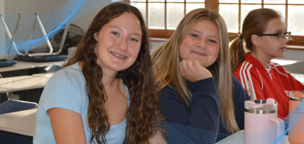 Three students seated at desks in classroom. Two are smiling for the camera on first day of school