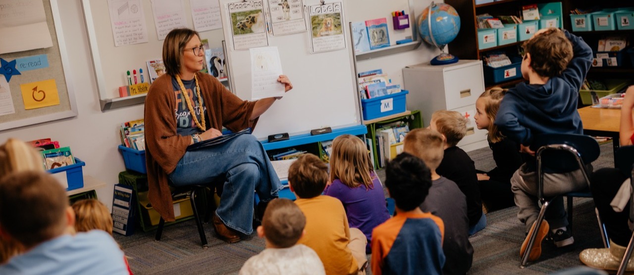 Teacher teaching seated children in classroom. Whiteboard and globe behind teacher. Children seated on floor in front of teacher.