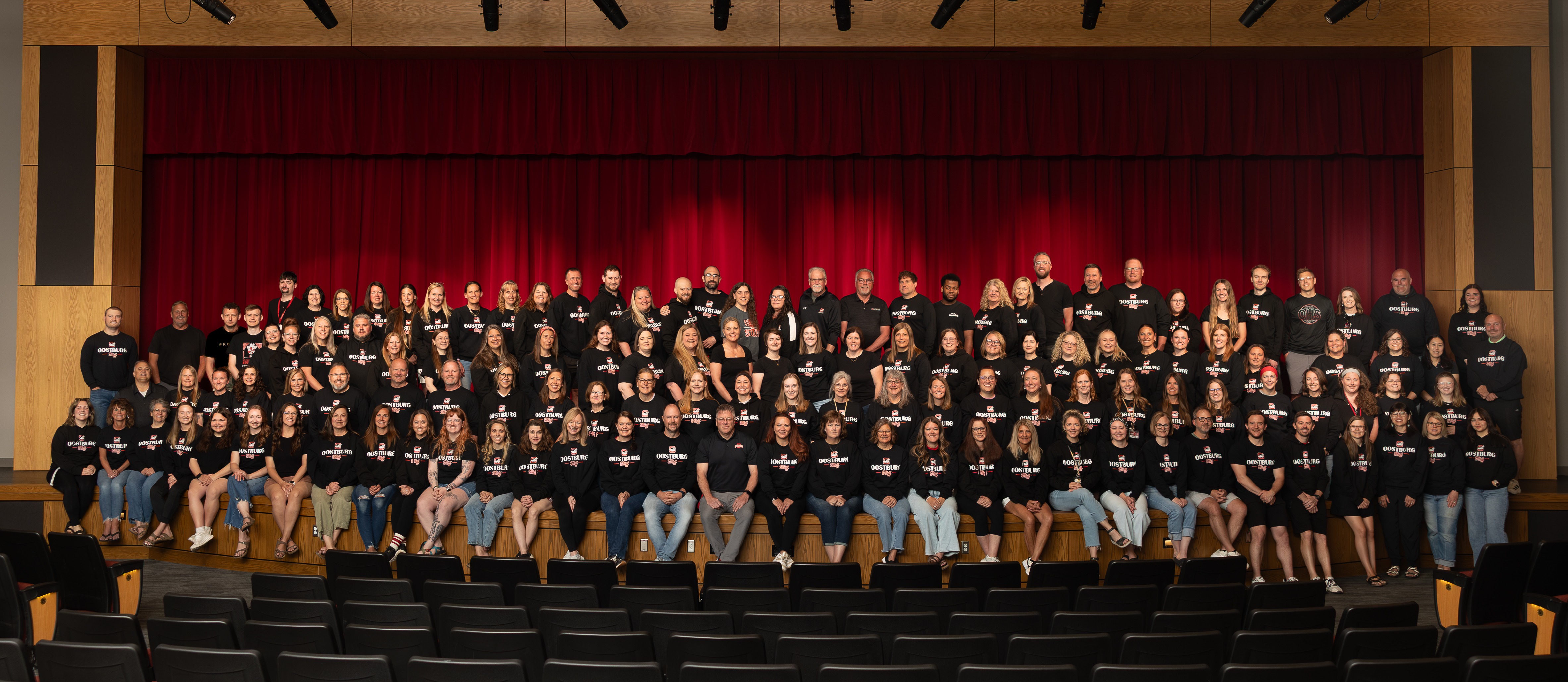 A group of people dressed in black shirts pose for a photo on a theater stage with a red curtain.