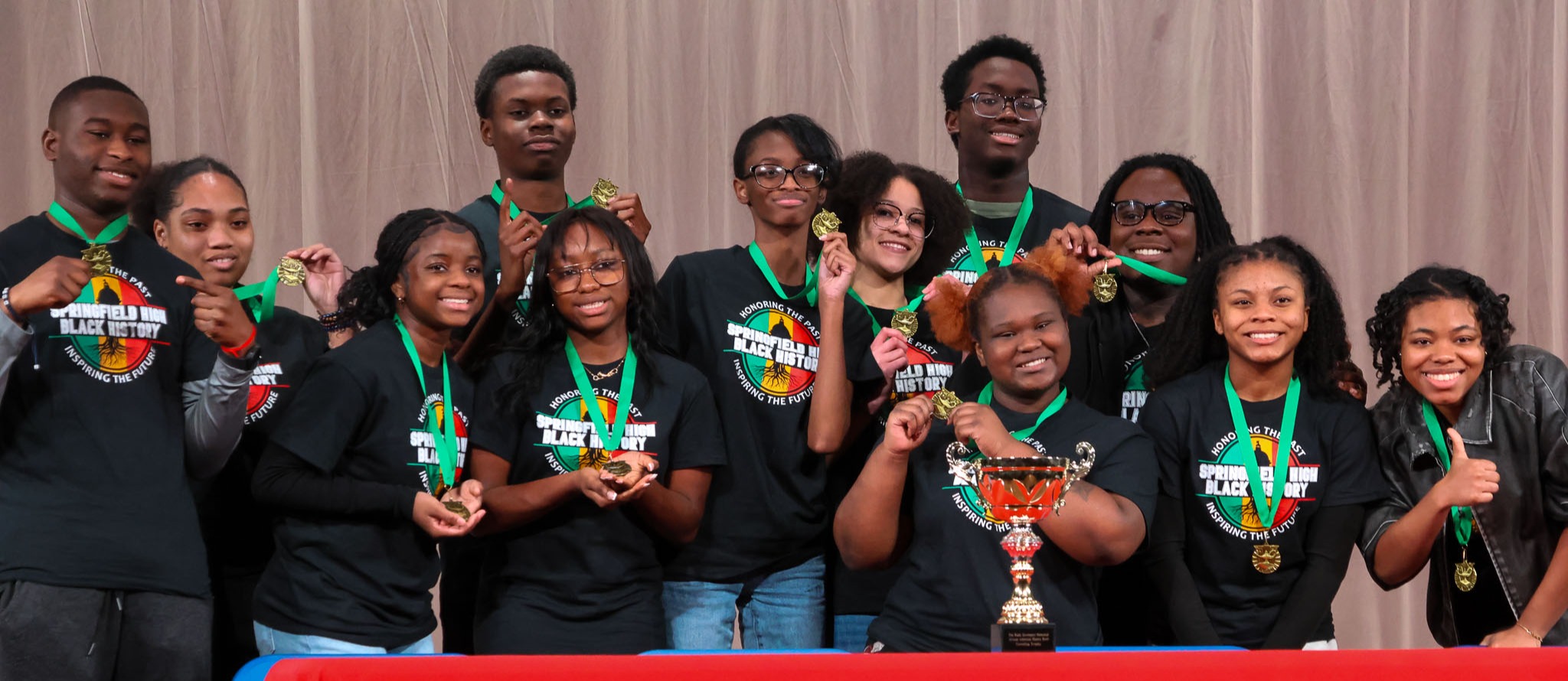 A group of people with medals stand behind a table with a red cover and a logo.