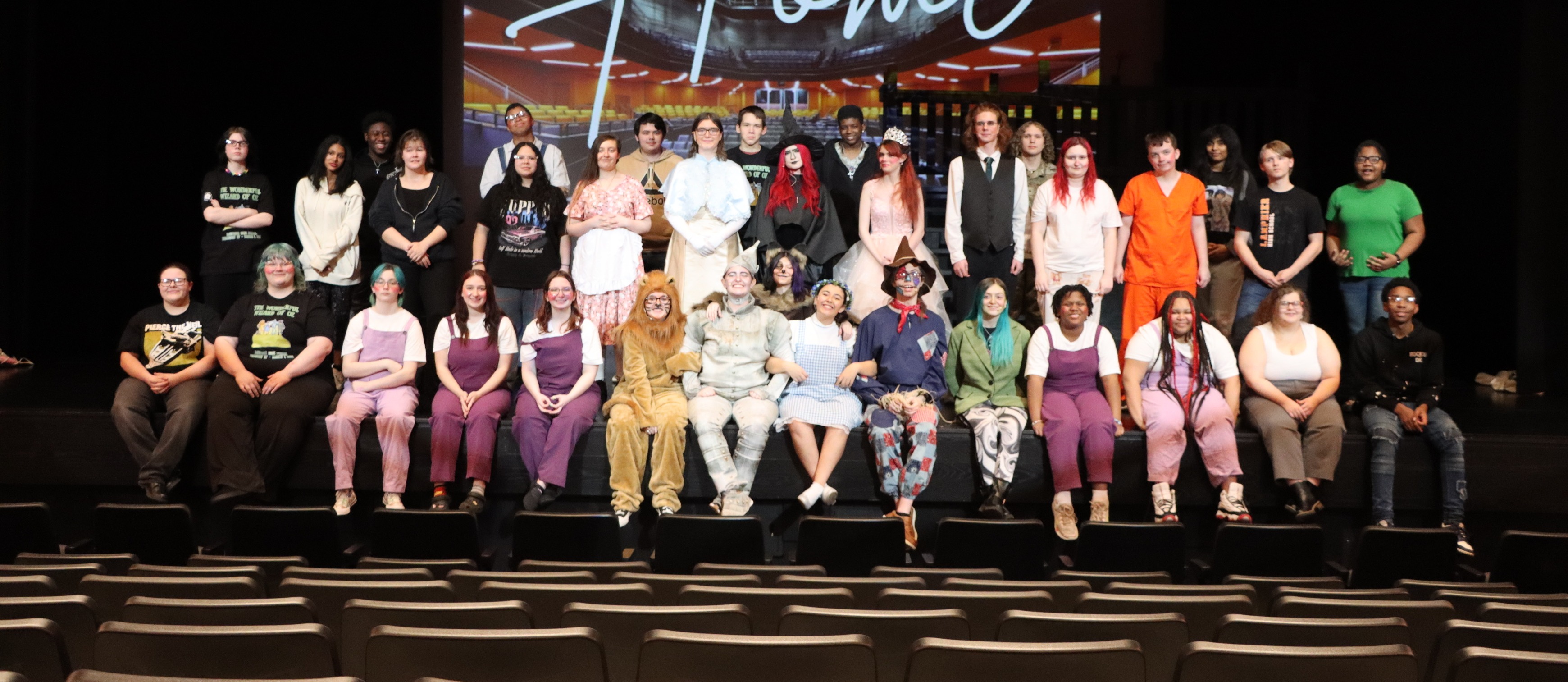 A group of people in various costumes stands on a stage with a large screen behind them.