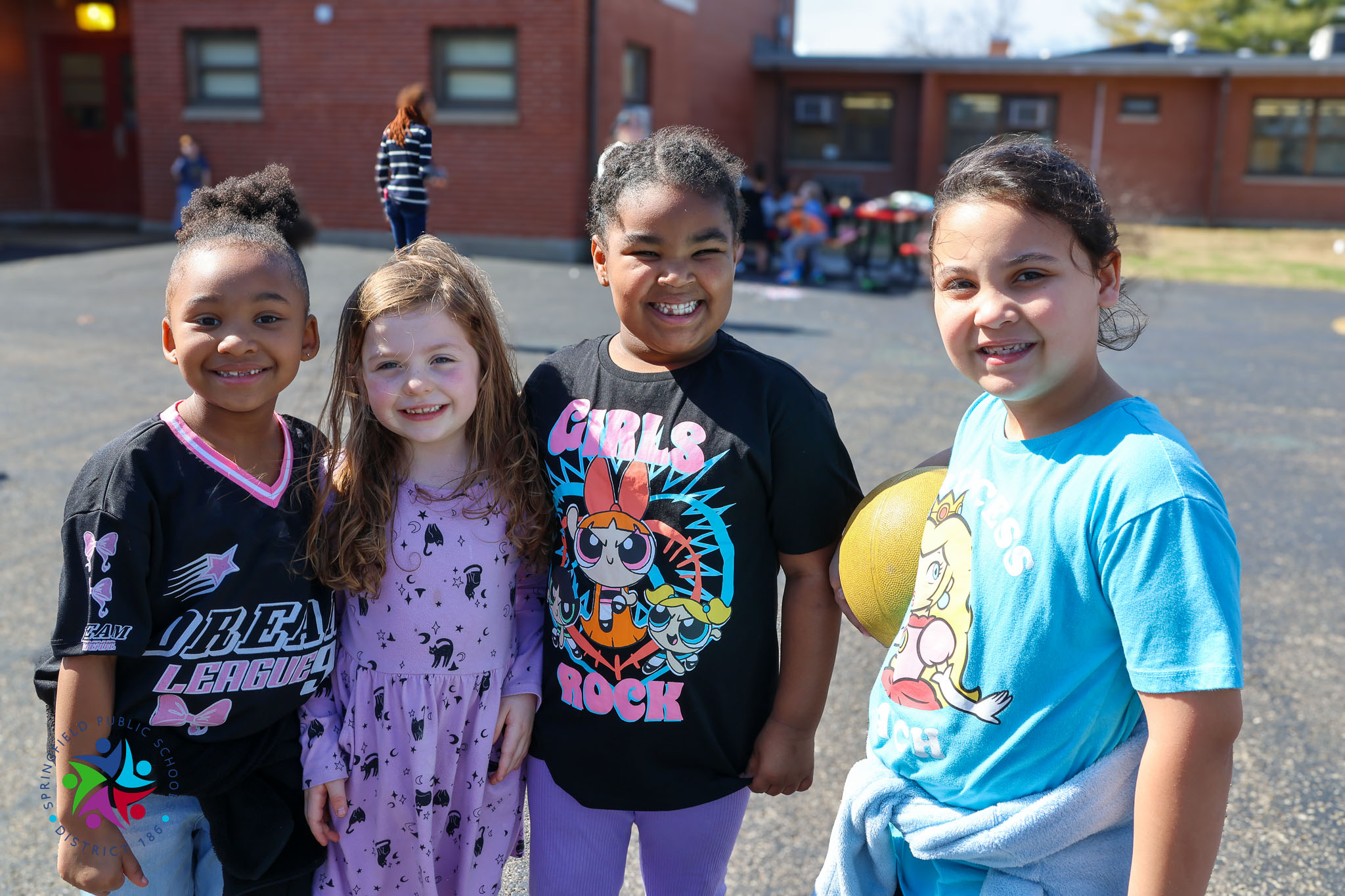 Four young girls stand together in a schoolyard, smiling. Behind them, a brick building is visible.