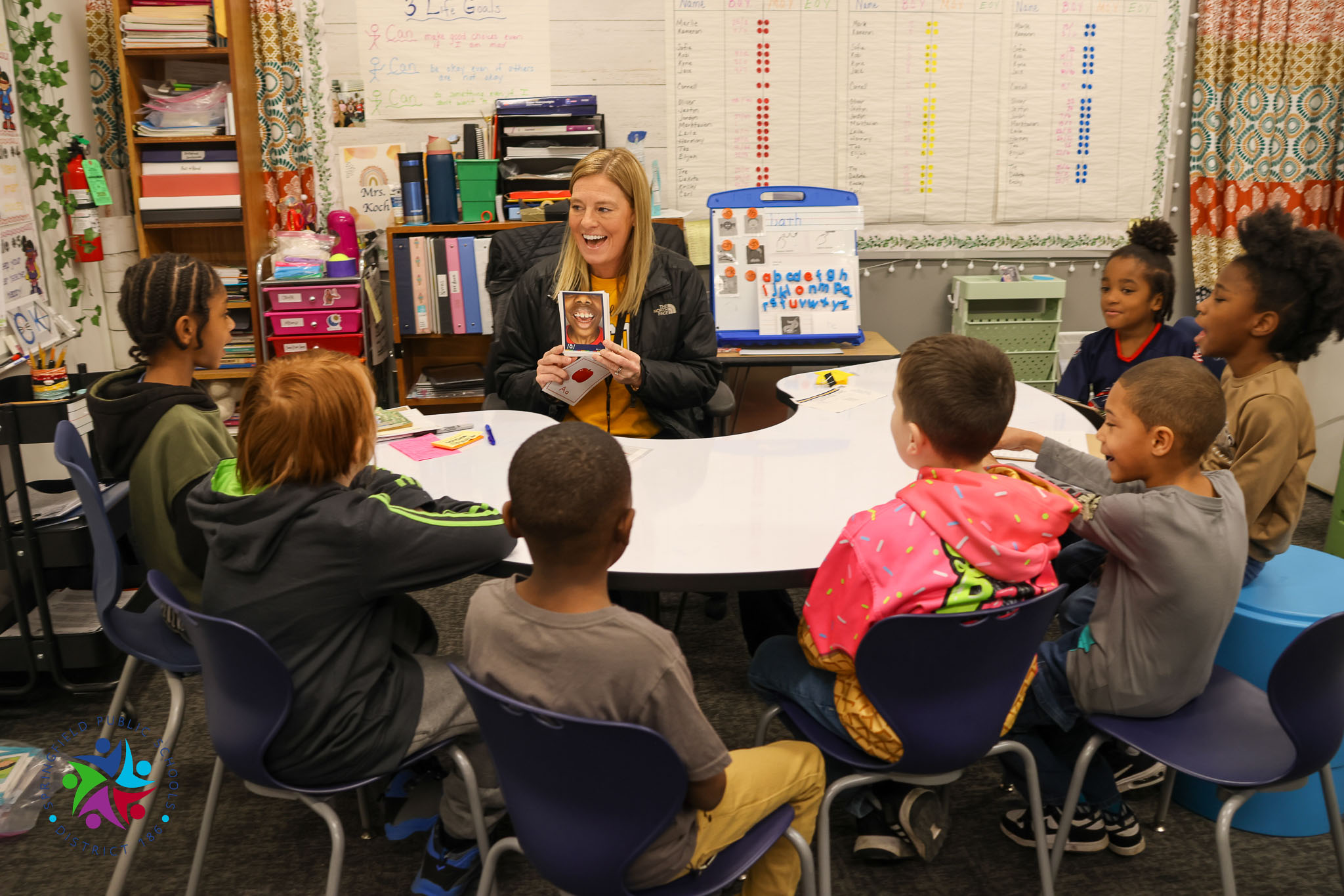 A group of children sit around a teacher in a classroom. The teacher holds a book and points at it.