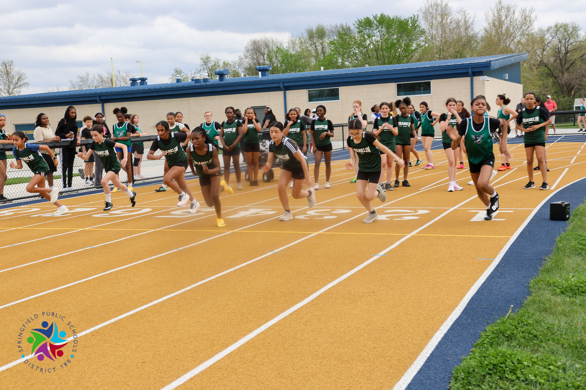 Participants in green uniforms run on a track with white lines. Some are at the starting line, while others are in motion.