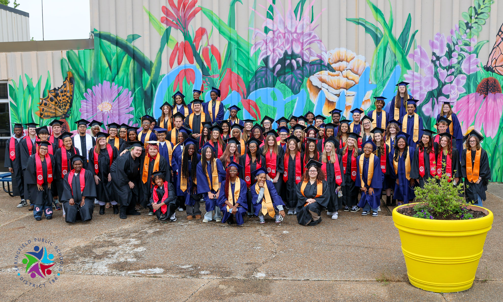 Graduates in caps and gowns pose for a photo in front of a colorful floral mural.