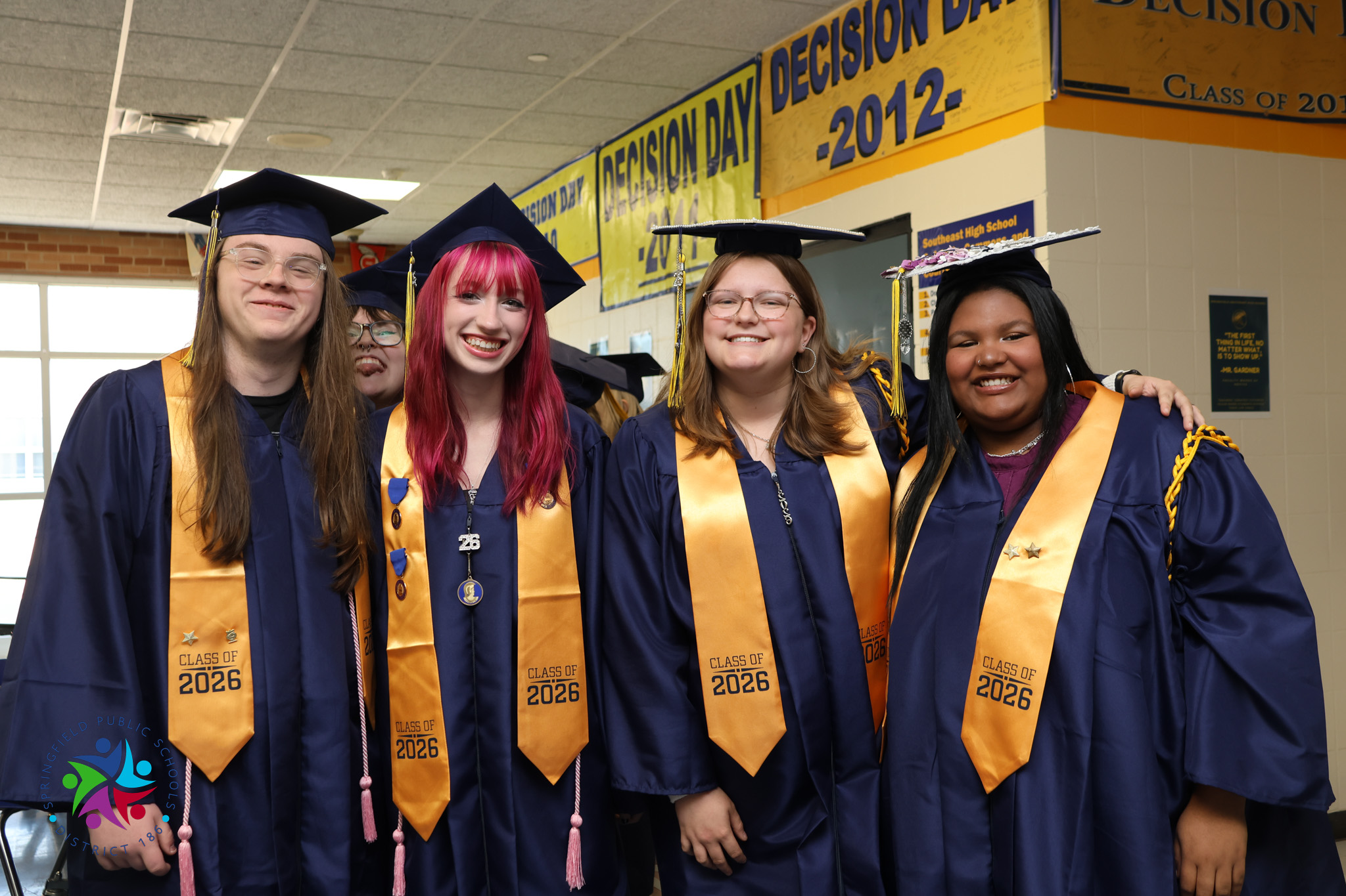 Five individuals in graduation robes and caps stand together. The two in front wear red hair and glasses.