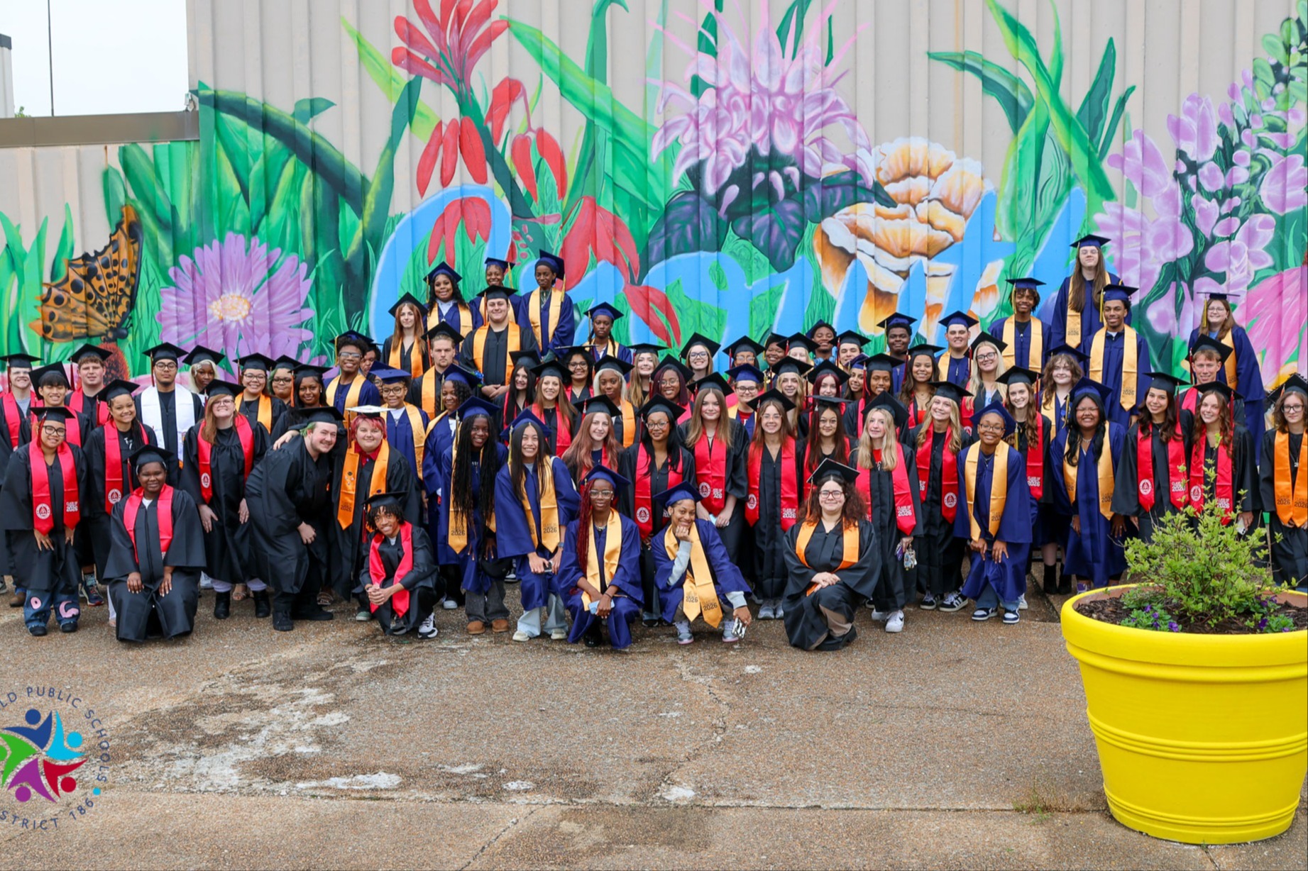 Graduates in caps and gowns pose in front of a building with a colorful mural.