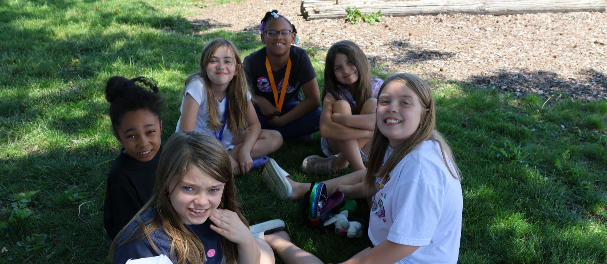 A group of girls sit on grass. They are smiling and holding papers. One wears a white shirt.