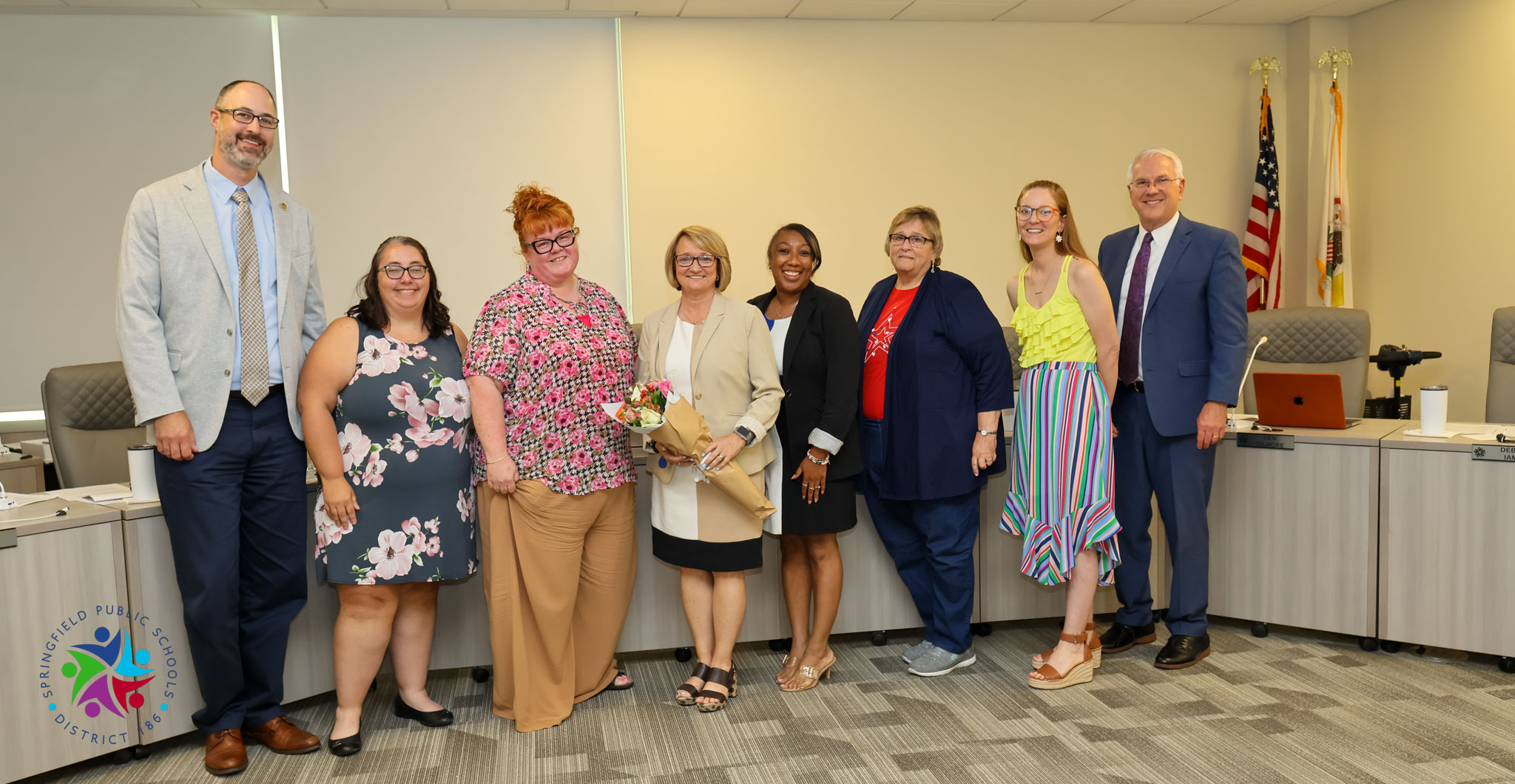 Seven people stand in a line in an office. One holds flowers. Flags are on poles behind.