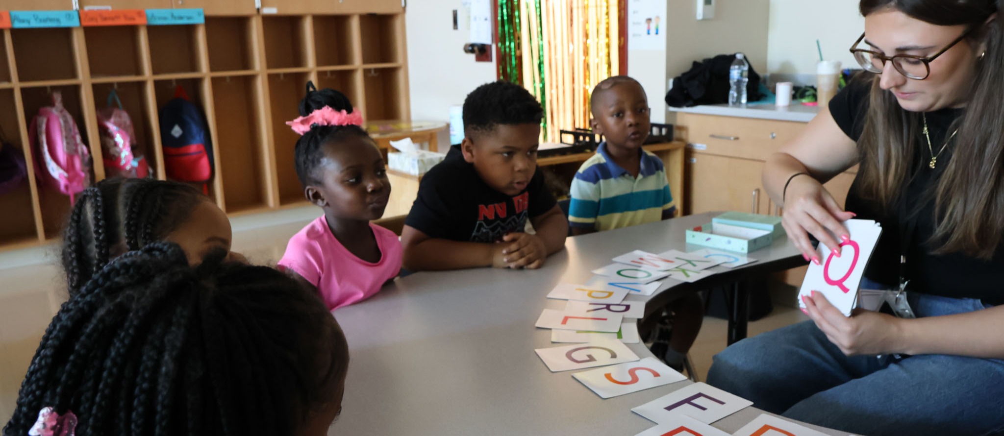 A woman sits with four children at a table with alphabet cards. The woman holds up a card.