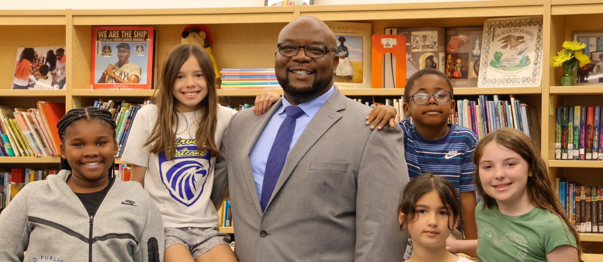 A man stands in a library with six children, all smiling, with shelves full of books in the background.
