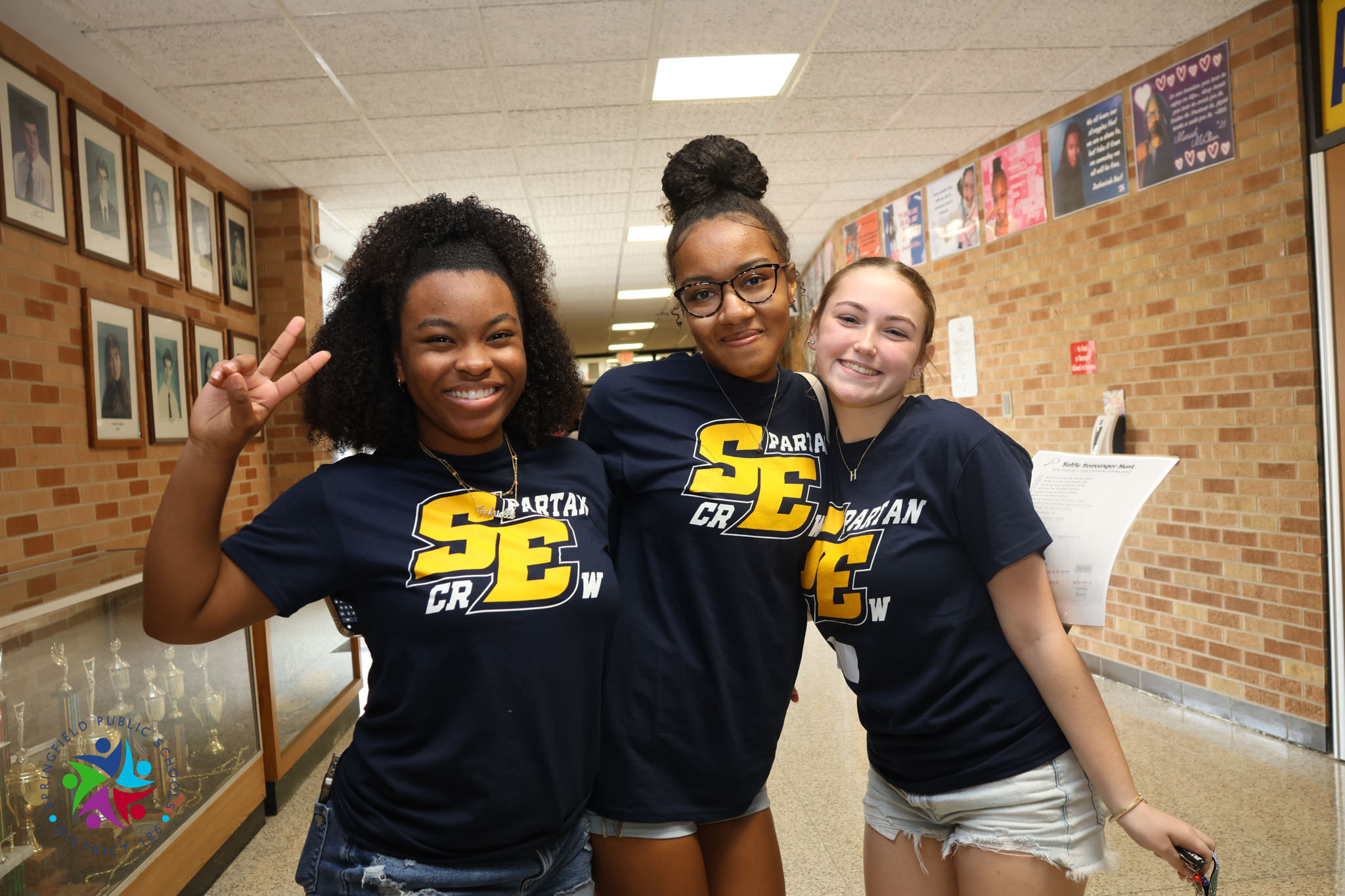 Three women wearing matching shirts stand in a hallway, smiling and posing for a photo.