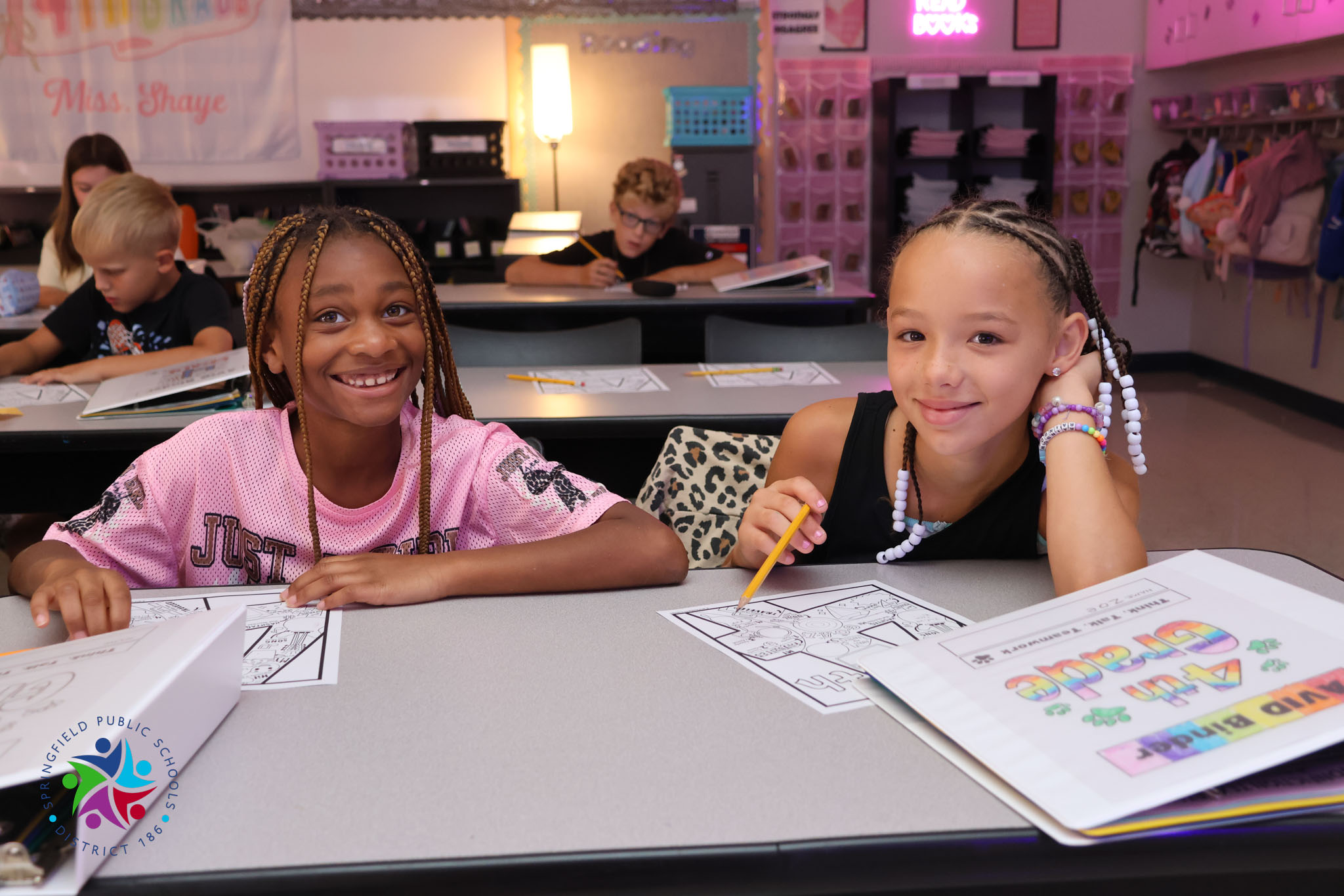 Two young girls seated at a table, smiling, with papers and pencils. Other children and adults are seated at tables in the background.
