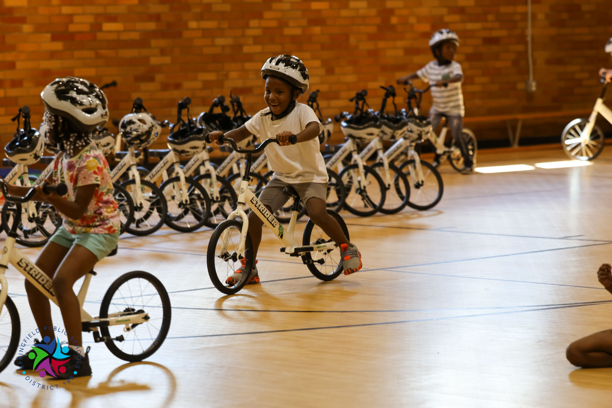 Children wearing helmets ride bicycles on a wooden floor in a room with brick walls.