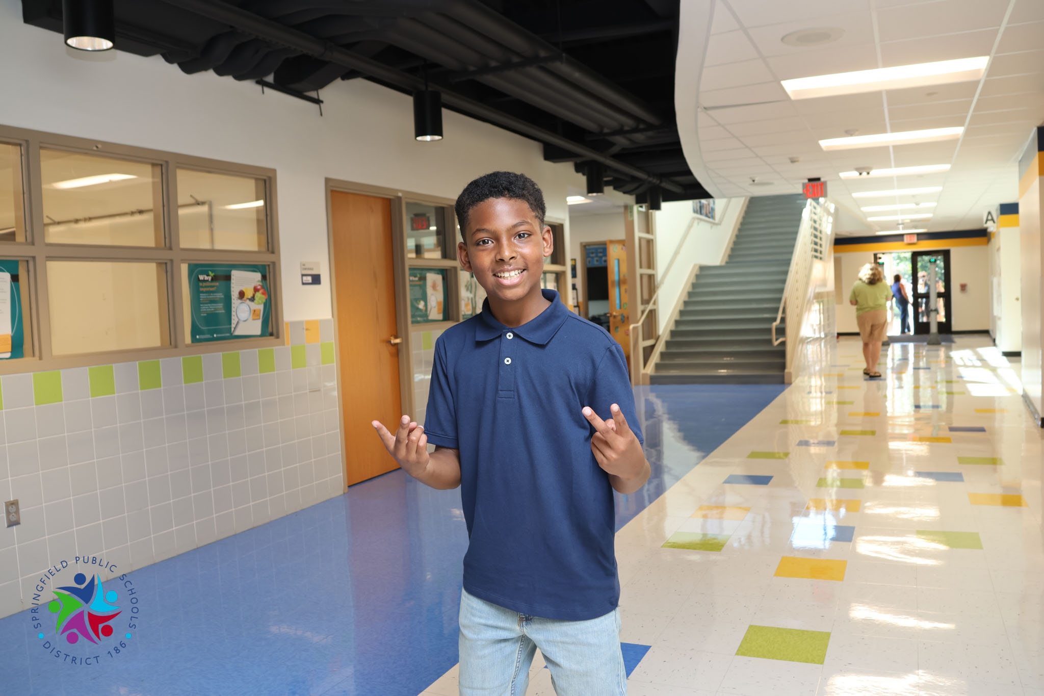 A young boy with a smile, wearing a blue polo and light blue jeans, stands in a school hallway.