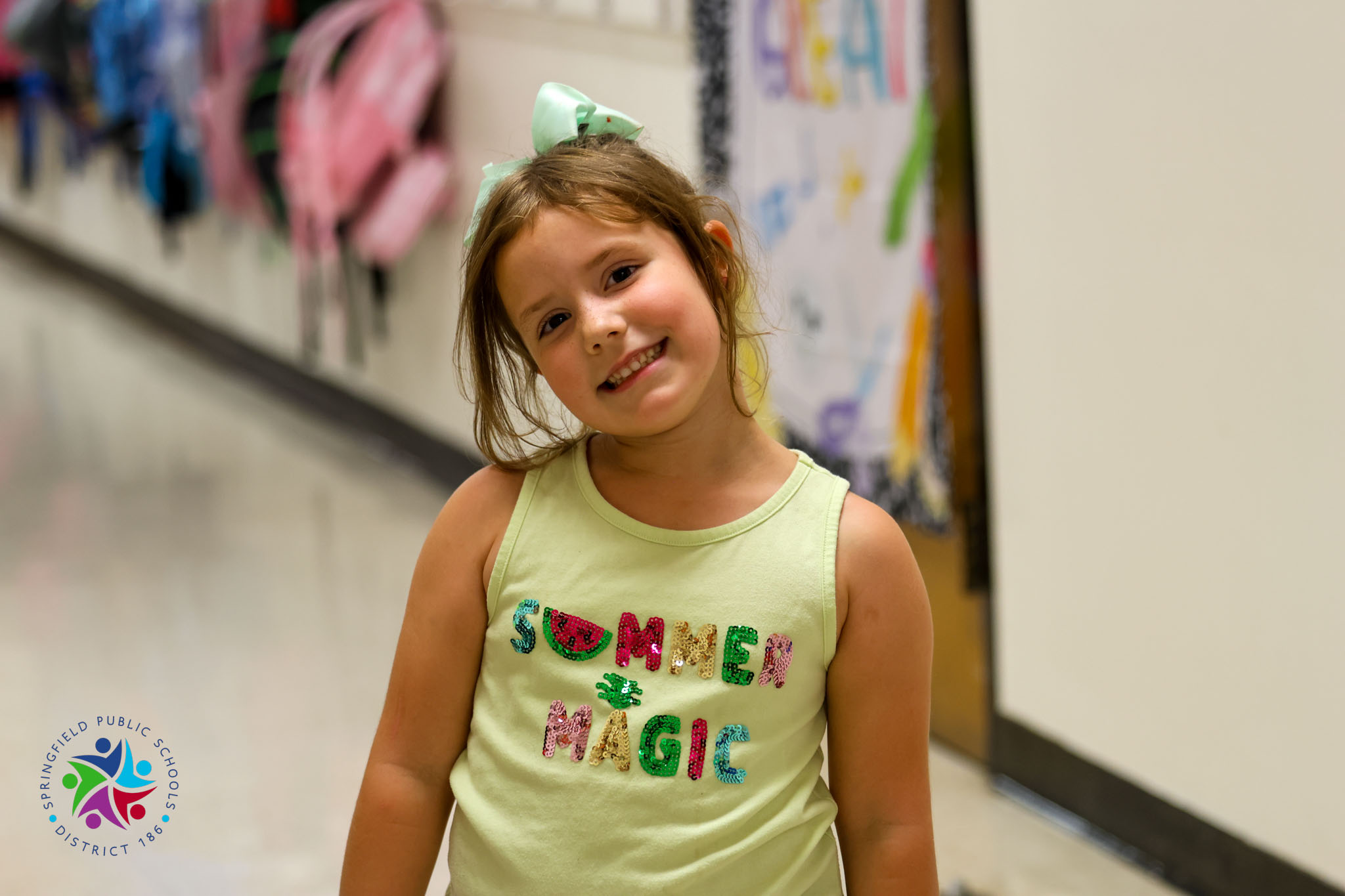 A girl with a green bow stands in a hallway. She wears a yellow shirt with "SUMMER MAGIC" on it.