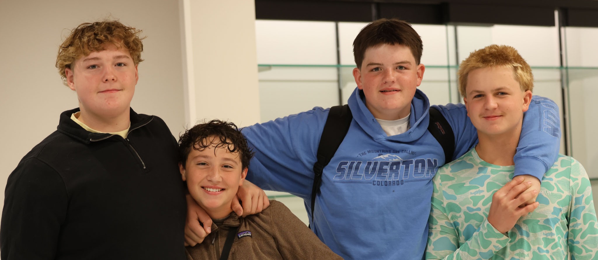 Four young men stand together, smiling. They wear various jackets, and one holds books. A white wall and a glass door are in the background.