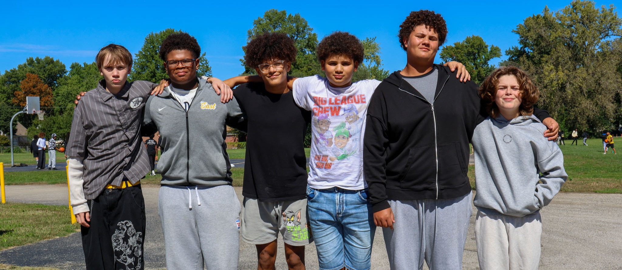Six young people stand in a row in a paved area with trees. They wear various colors of jackets and pants.