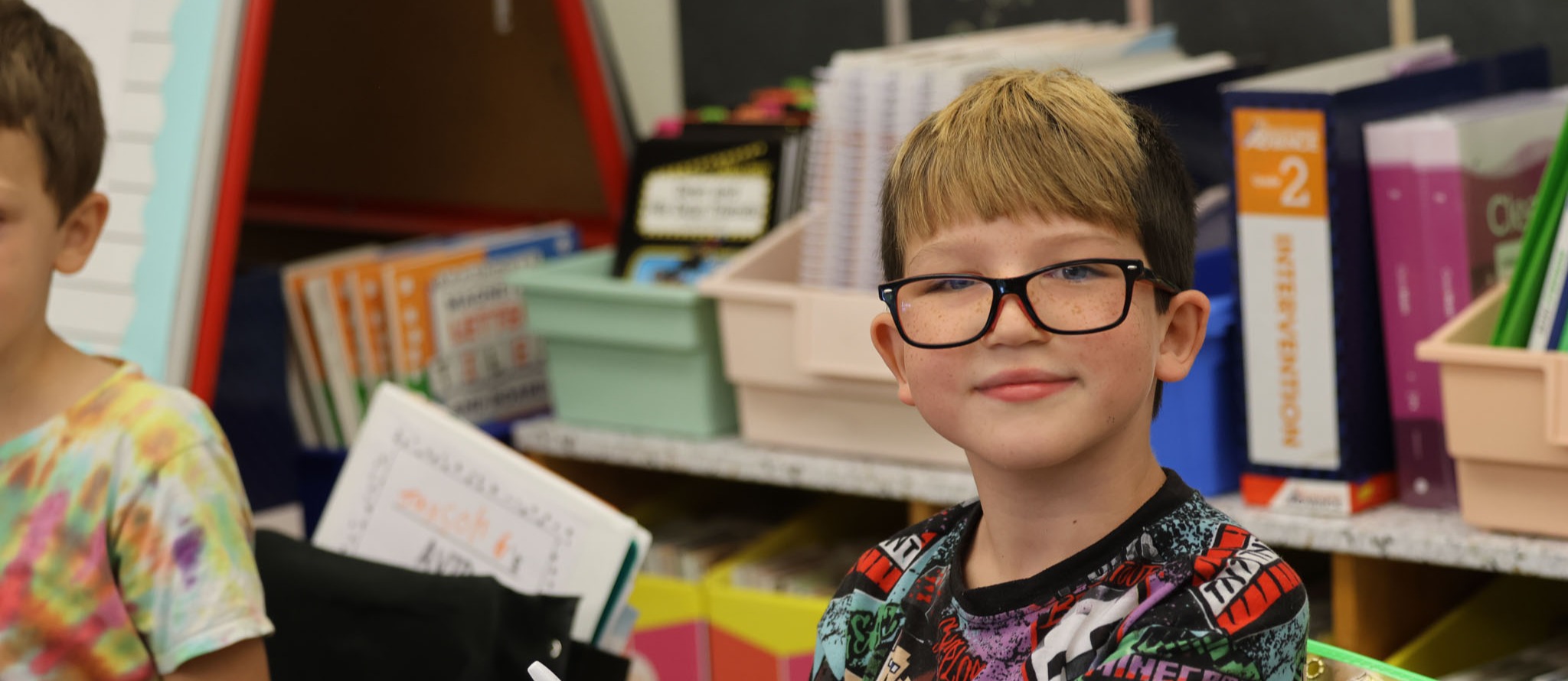 A boy with glasses sits at a desk, holding a pen and smiling. Another boy in a tie-dye shirt sits behind him.