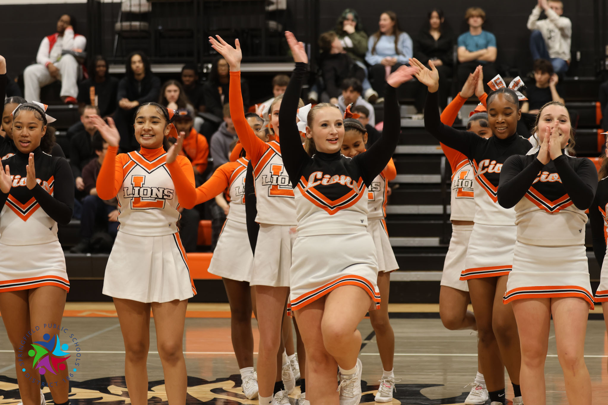 Seven cheerleaders in matching uniforms clap hands in front of a cheering crowd on a court.