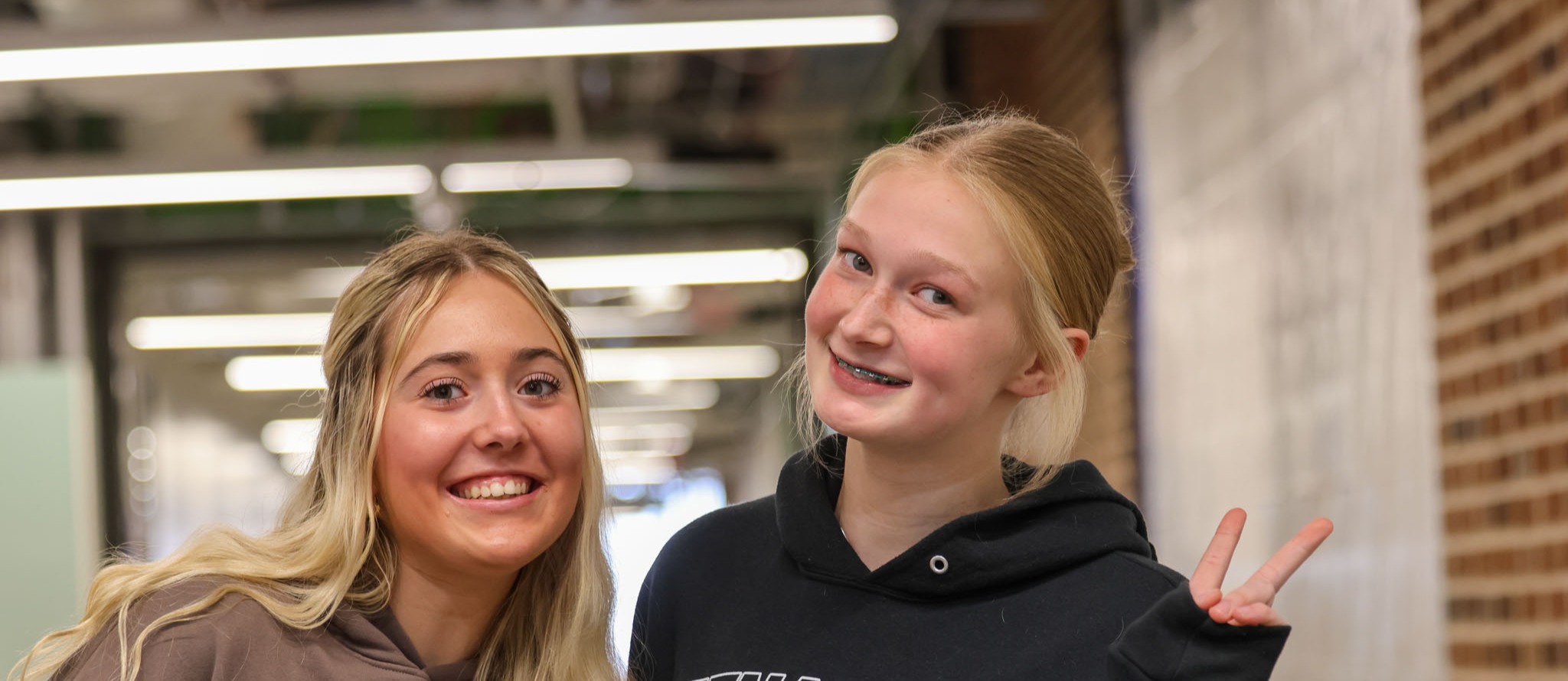 Two girls stand in a hallway, smiling, one with blonde hair, the other in a black hoodie with volleyball text.