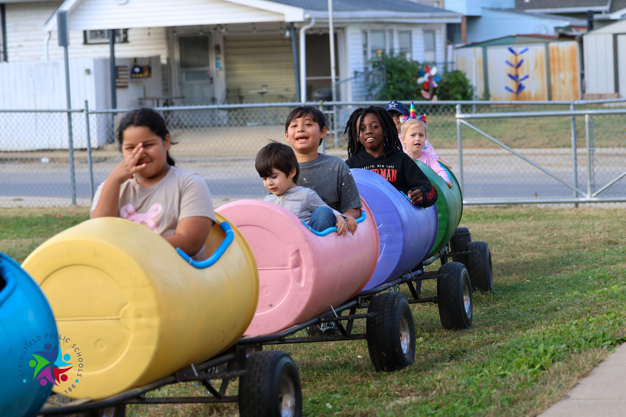 Children in colorful tubes ride a small cart on a grassy area, with houses in the background.