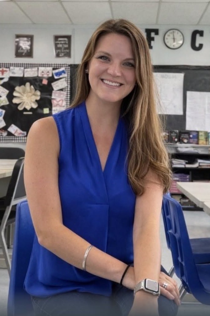 A woman in a blue blouse sits in a classroom with tables and chairs behind her.