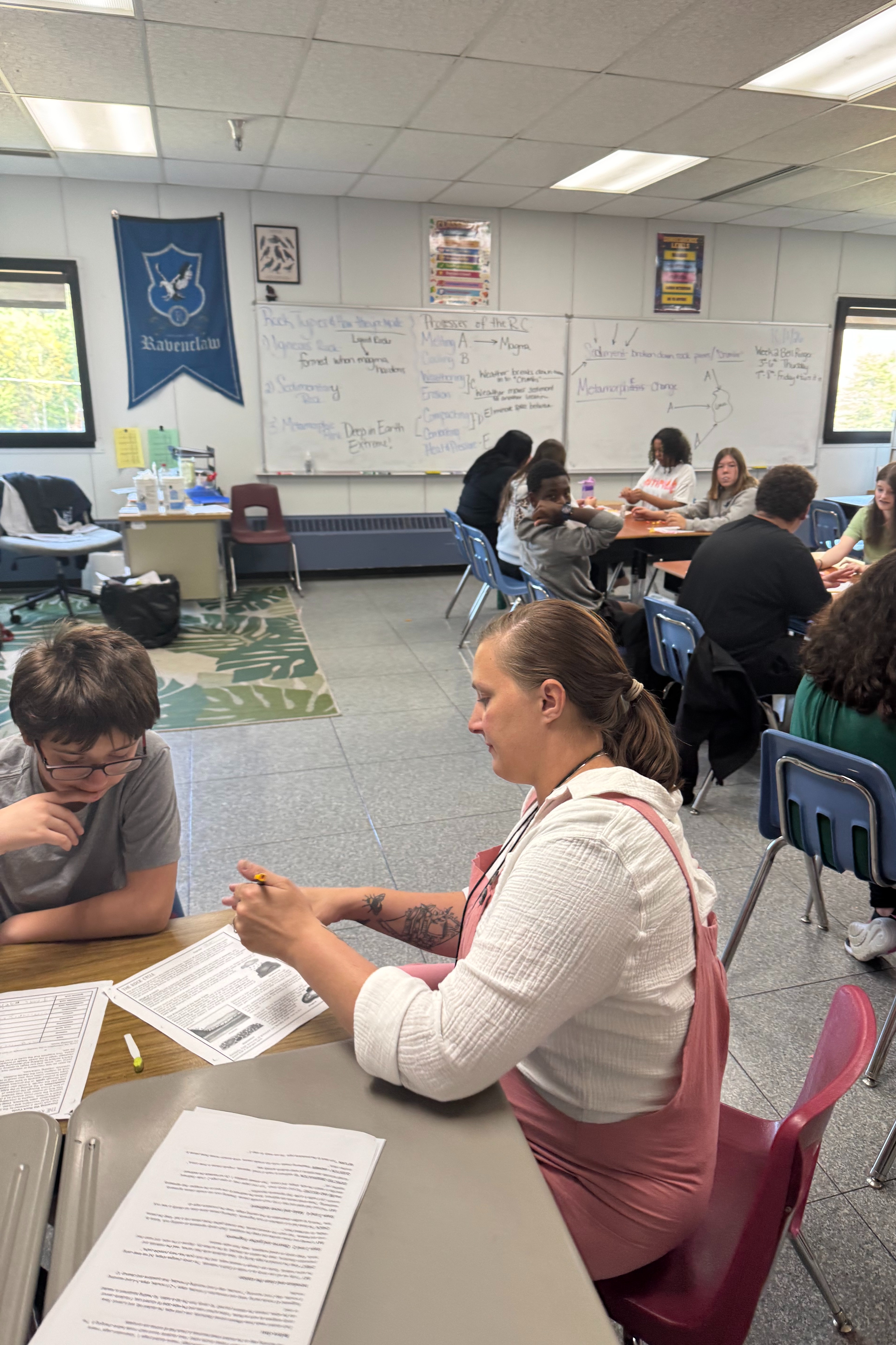 A classroom with students seated in chairs. A teacher assists a student. Papers and books on a table.