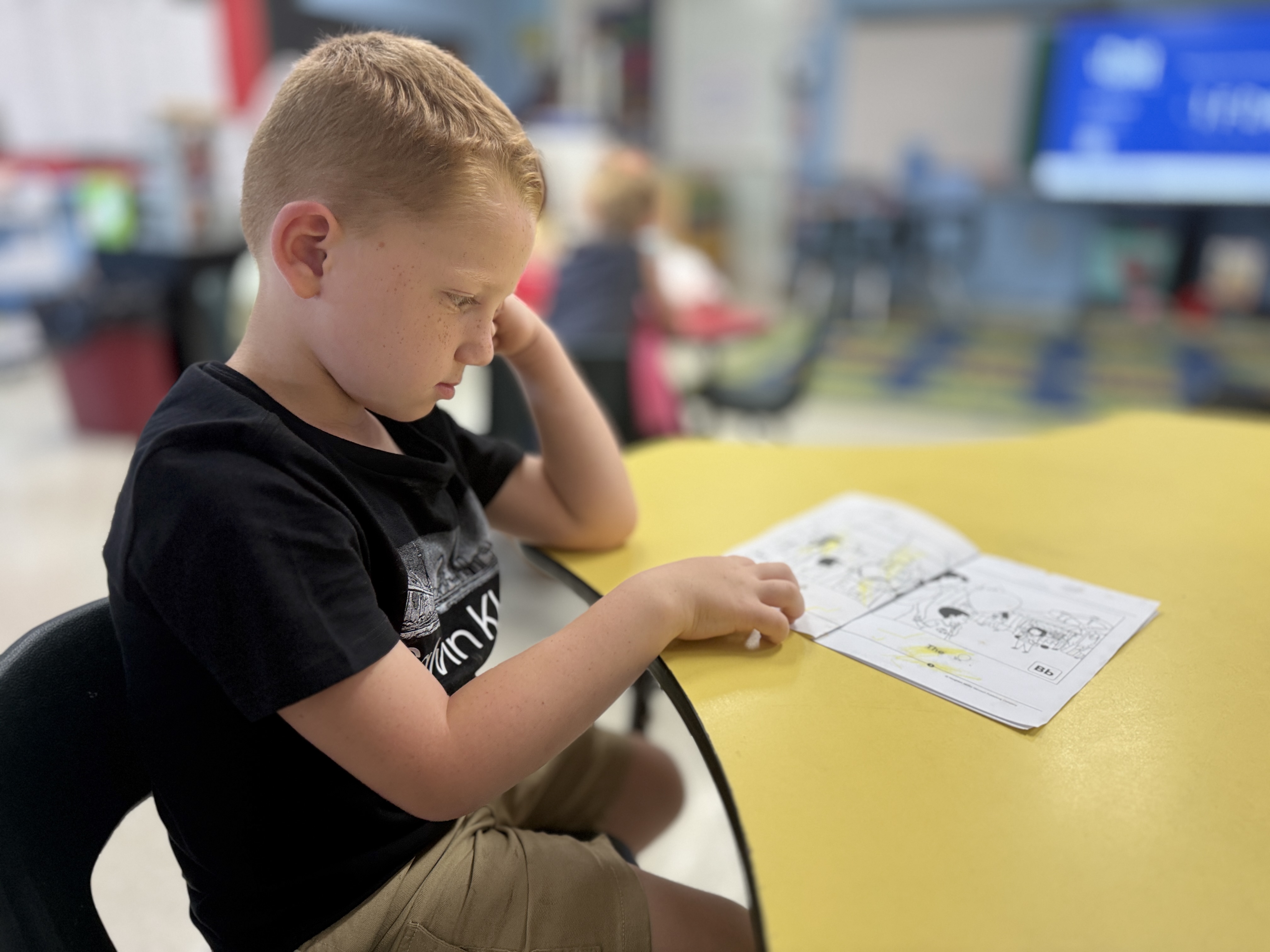 A young boy in a black T-shirt and khaki shorts sits at a yellow table, studying a white sheet of paper.