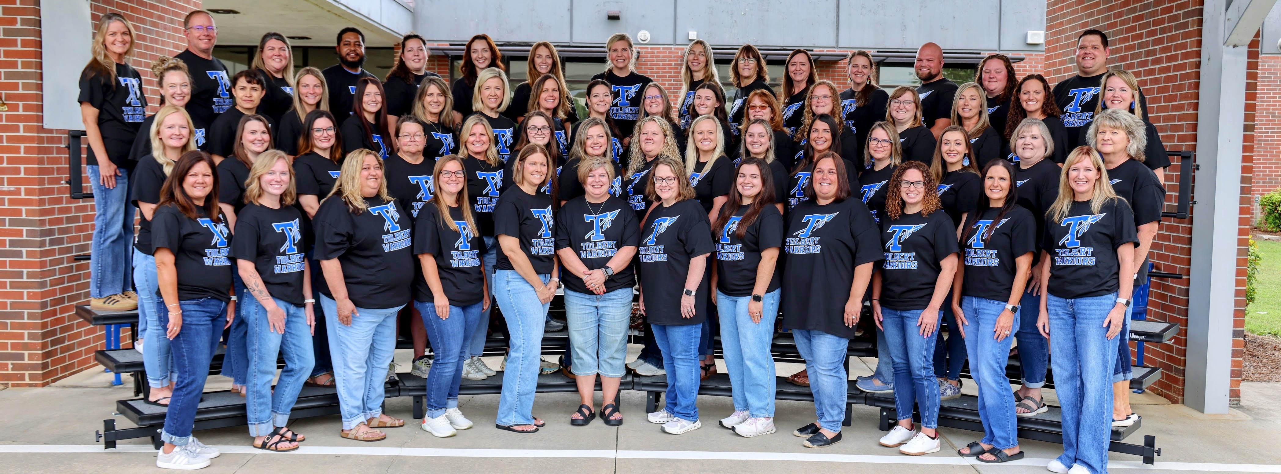 Tolbert Elementary School faculty and staff pose together on risers outside the school entrance wearing matching black Tolbert Warriors T-shirts.