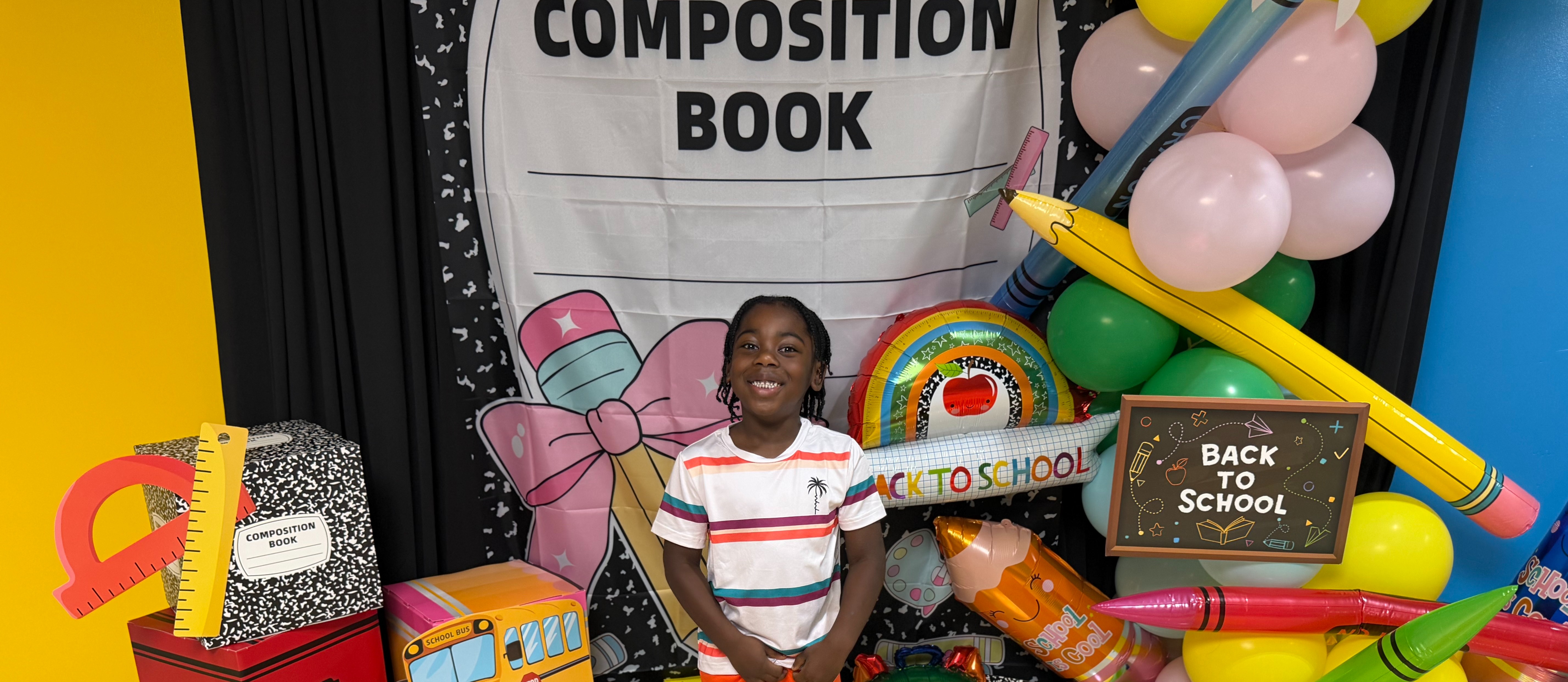 A child stands in front of a colorful display of school supplies and balloons, with a banner reading "COMPOSITION BOOK".