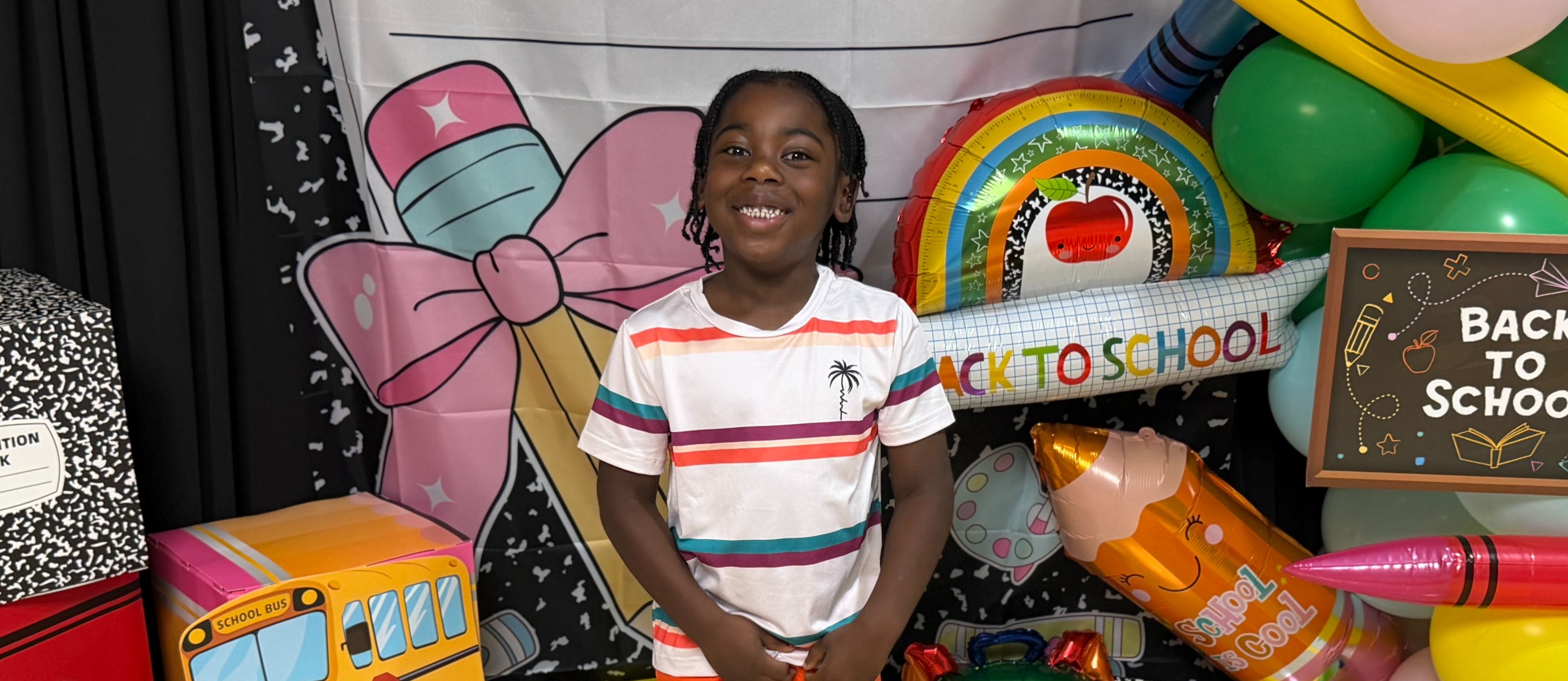 A child stands in front of a colorful display of school supplies and balloons, with a banner reading "COMPOSITION BOOK".