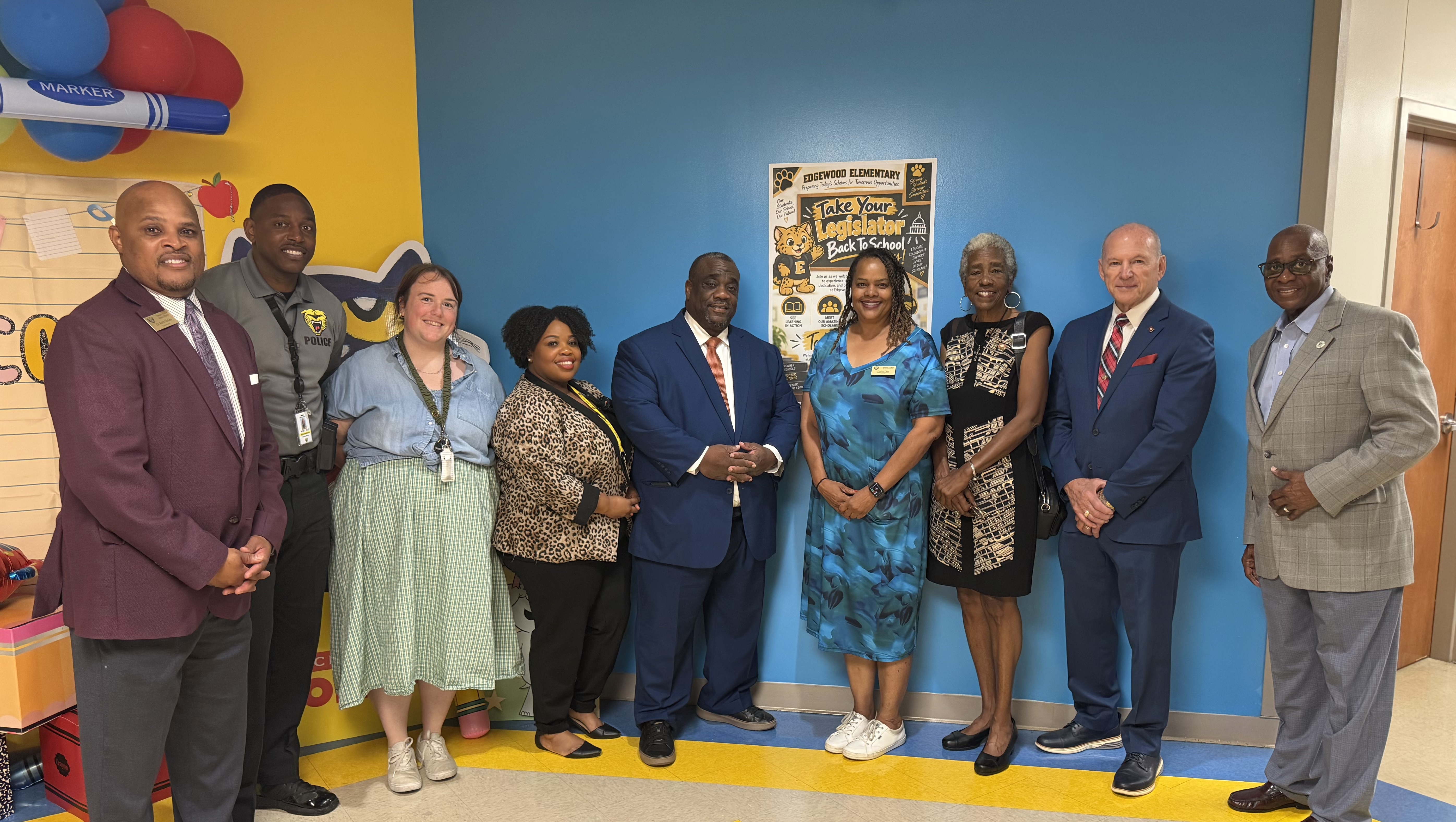 Nine people stand in a room with blue walls and yellow floor. They are dressed in formal attire.