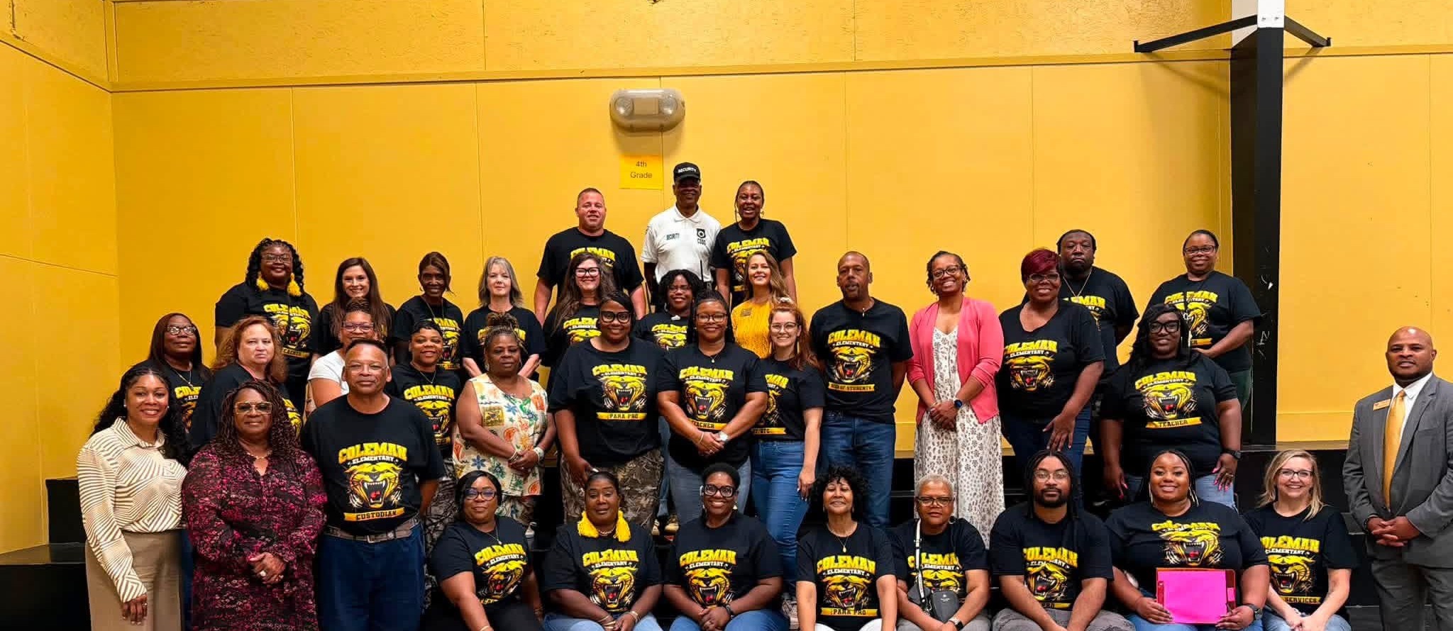 A group of adults and children pose for a photo in a gymnasium with yellow walls.