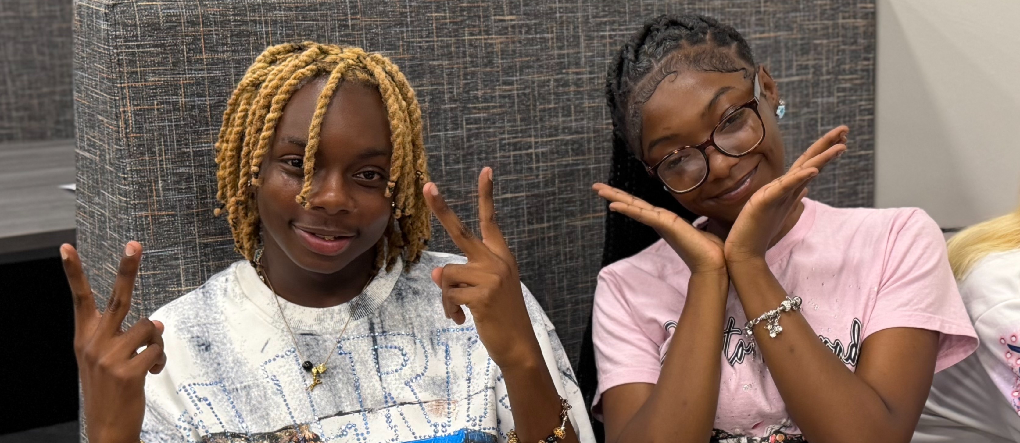 Two individuals seated on a bench, one with blonde hair, both wearing glasses, making peace signs.