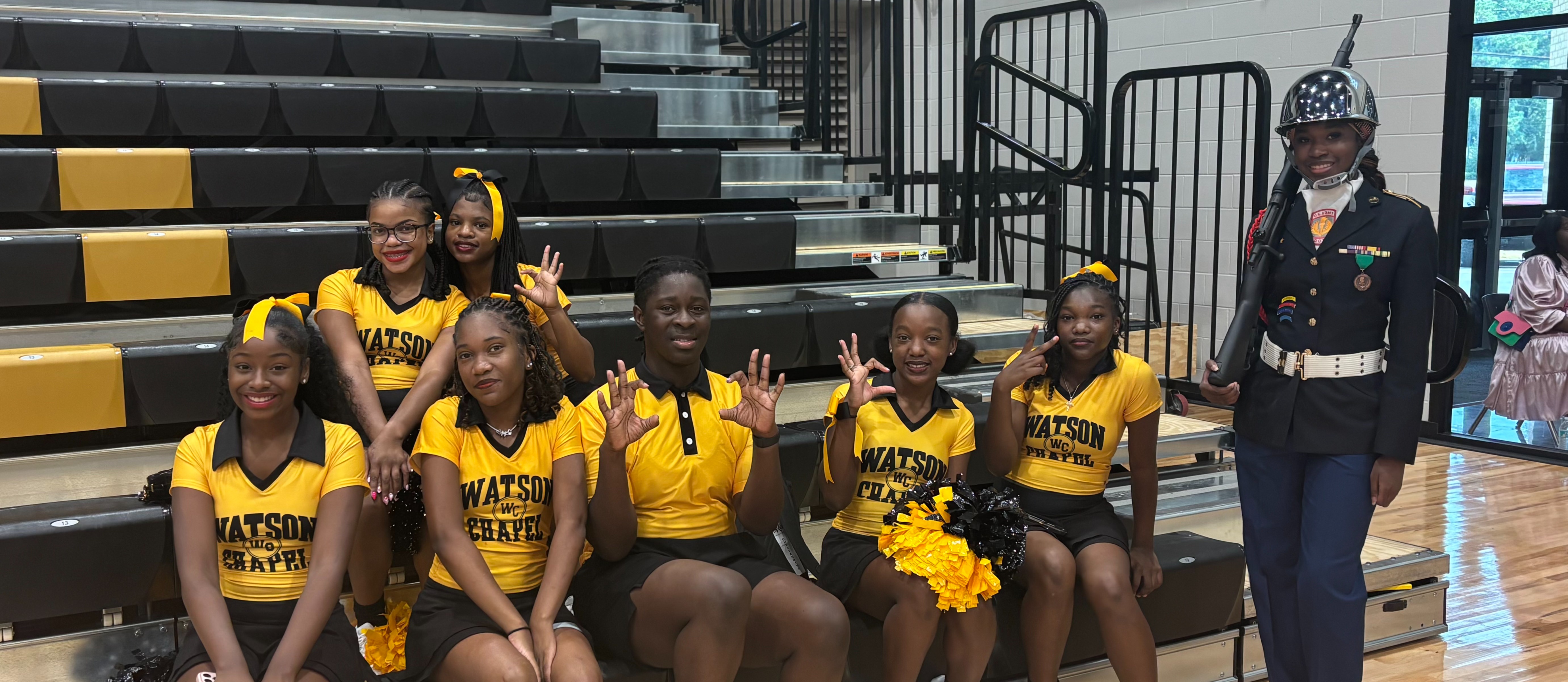 A group of cheerleaders in yellow uniforms sits on bleachers with a person in a uniform standing behind them.