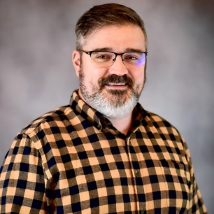 A male instructor wearing glasses and a yellow plaid-printed shirt smiles for the camera in front of a gray-colored backdrop.