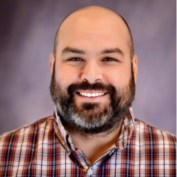 A male instructor smiles for the camera in front of a gray backdrop. He is wearing a red, black, and white plaid pattern shirt.
