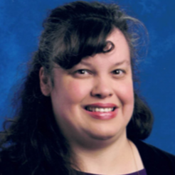 Dr. Christine Lewis. A female with dark curly hair smiles for the camera in front of a blue background. She is wearing a purple shirt with a black cardigan over top.