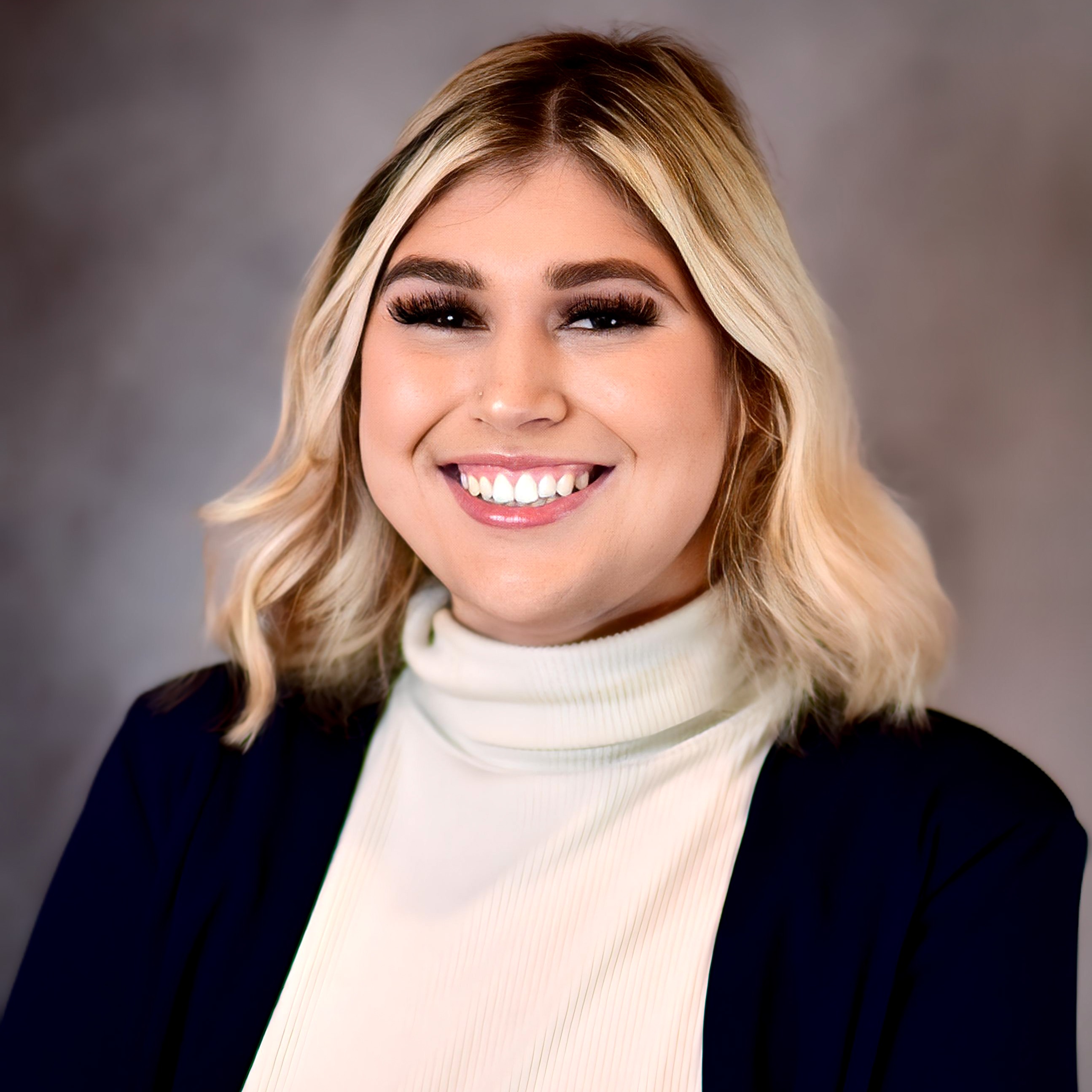 A female with blonde short hair smiles at the camera in front of a blue/gray backdrop. She is wearing a white turtleneck with a navy-colored blazer over top.