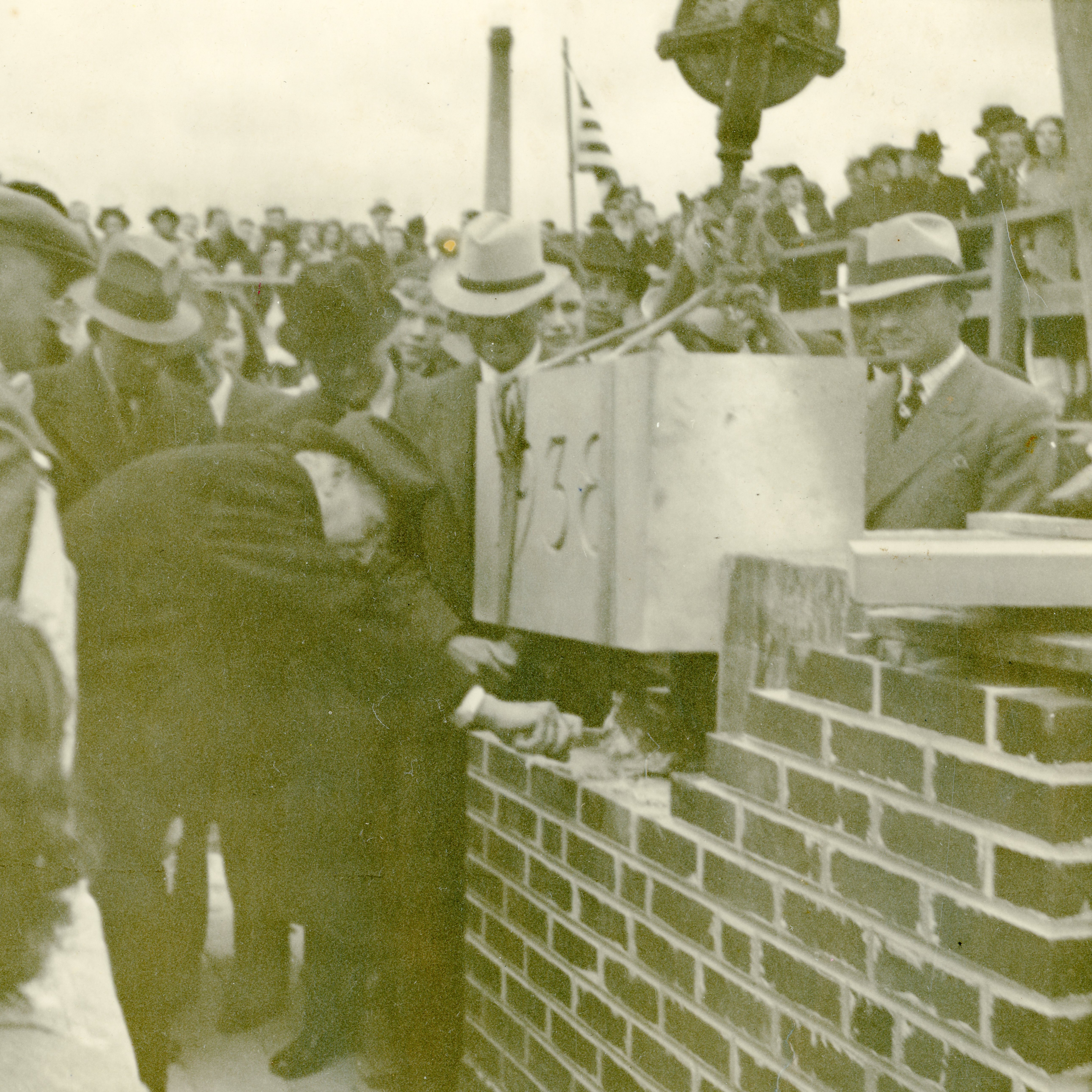Black and white photo of individuals placing a cement cornerstone onto brick with the date "1938" engraved into it.