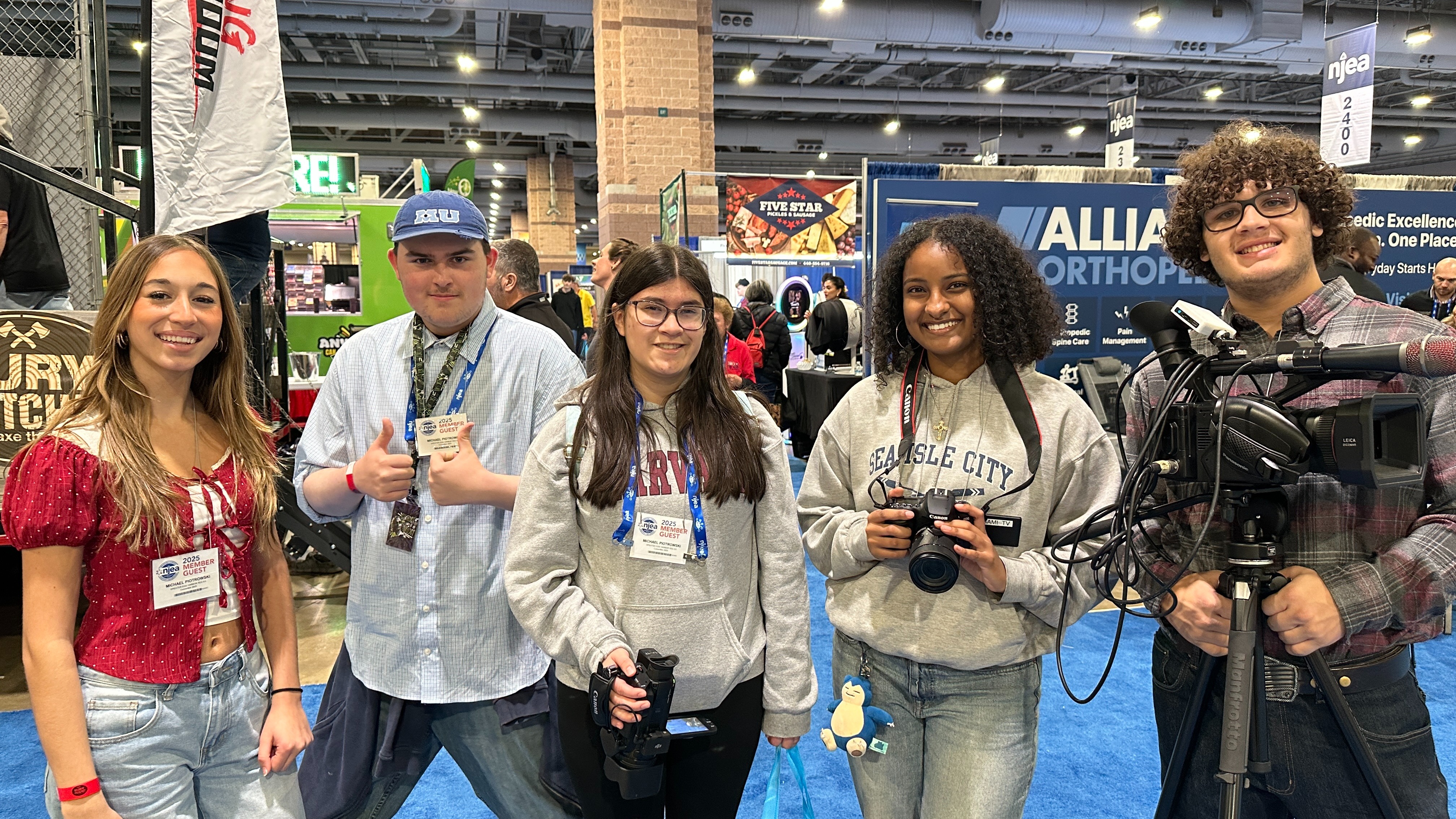 Five people stand together, smiling, at an event. One holds a video camera, while others hold cameras. A white banner with text is visible in the background.