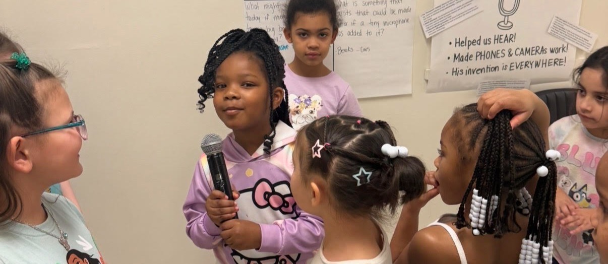 Group of children in a classroom. One holds a microphone, others look on. Background features posters.