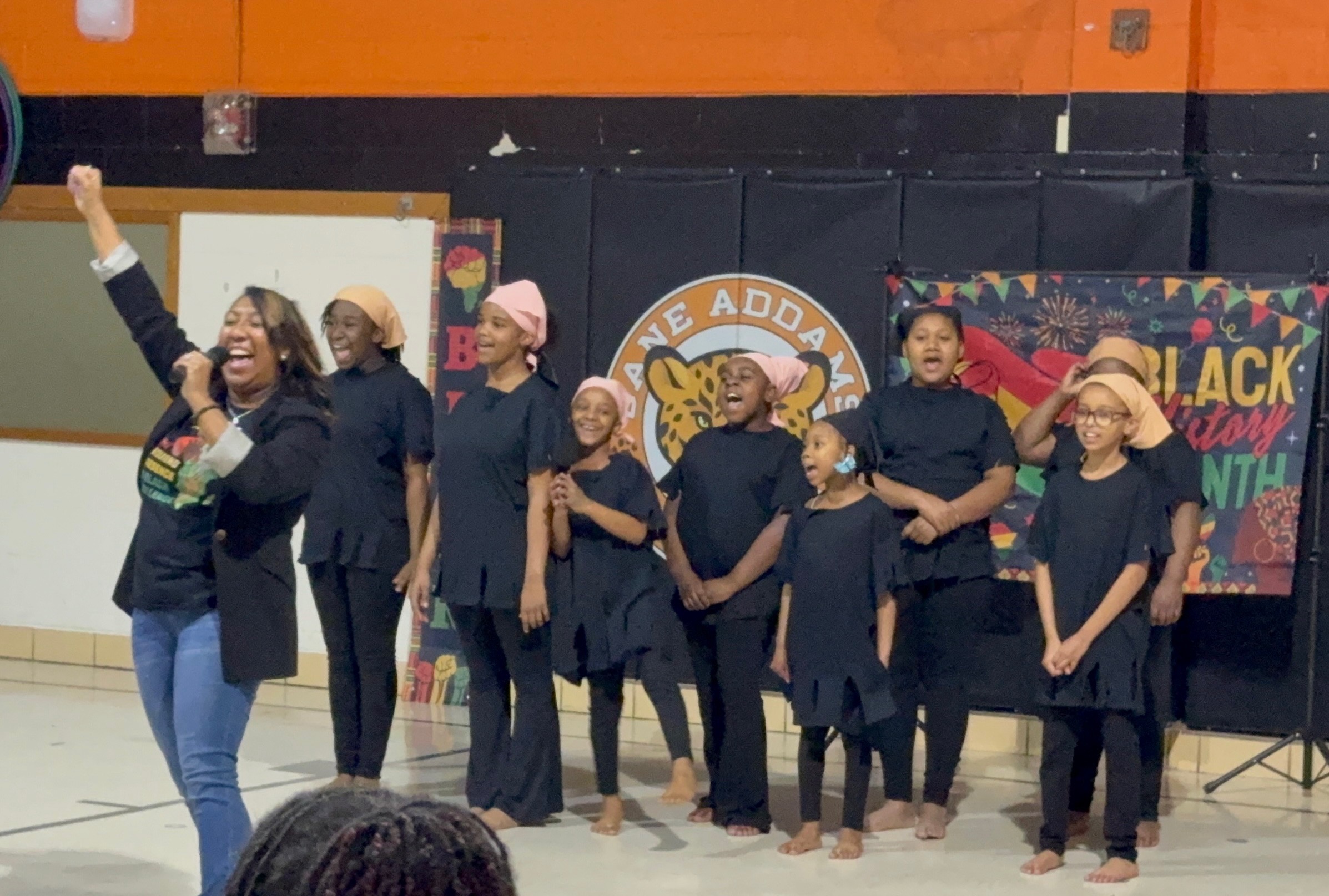 Group of children wearing black performing on stage with a woman standing in front of them.
